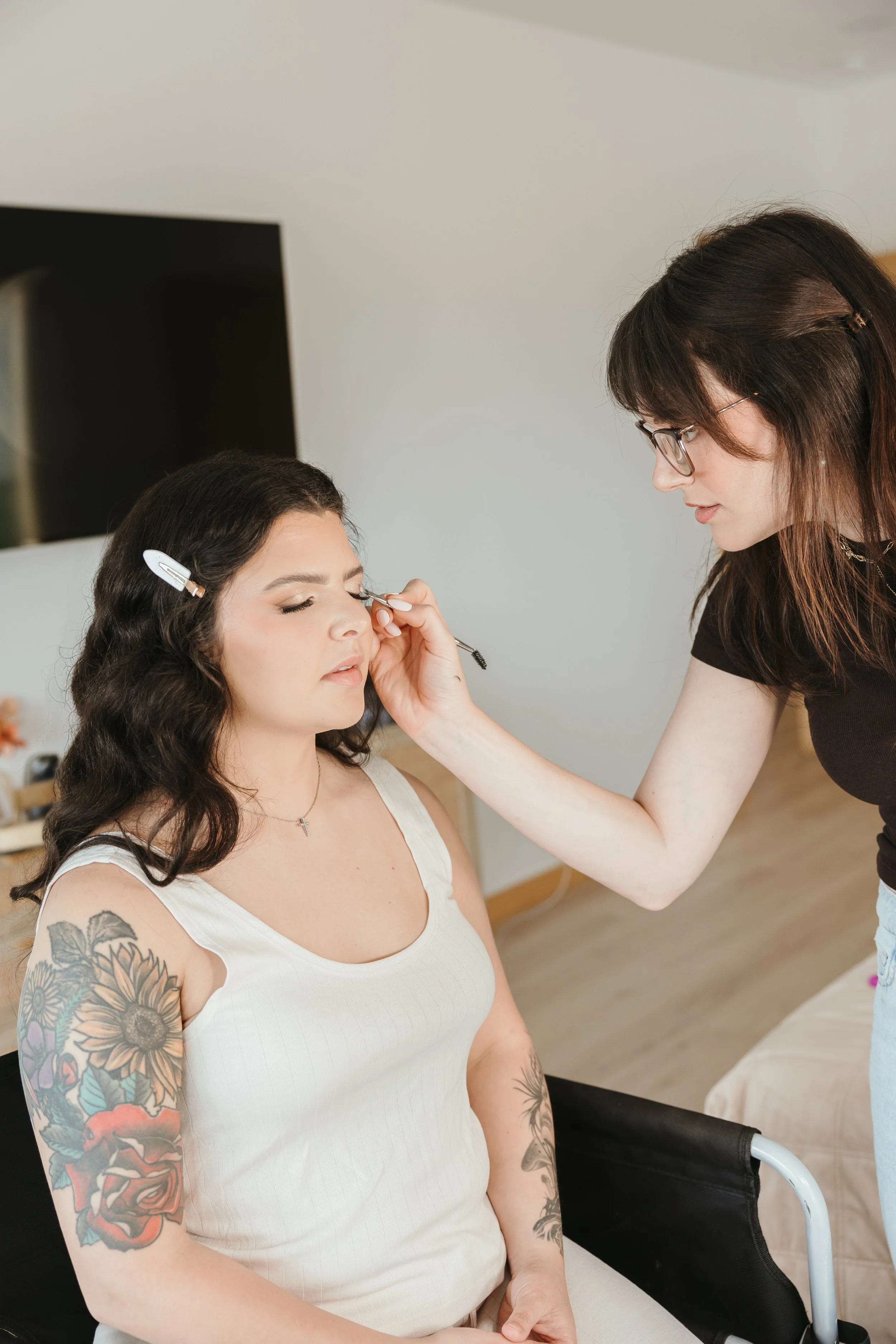 Makeup artist applying makeup to a woman with dark hair and tattoos on her arm, sitting in a chair.