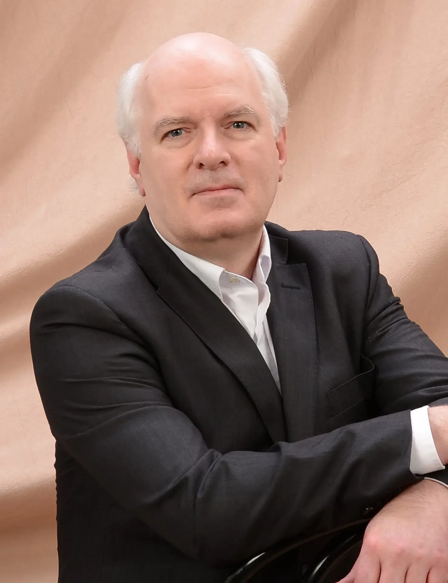 Headshot of Dr. Gregory Conn, a middle-aged man with white hair and a serious expression, wearing a black suit and white shirt, sitting against a beige background.