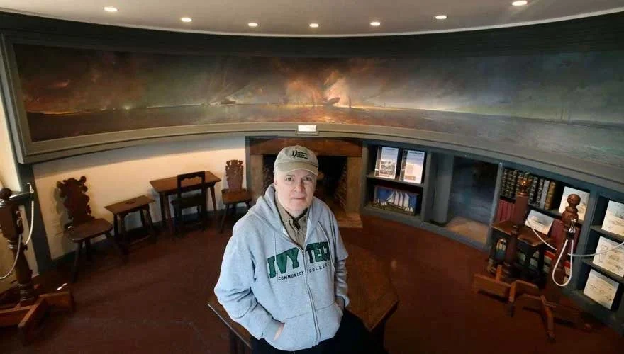 Man standing in a room with bookshelves and artwork, wearing a NYIT hoodie and baseball cap.