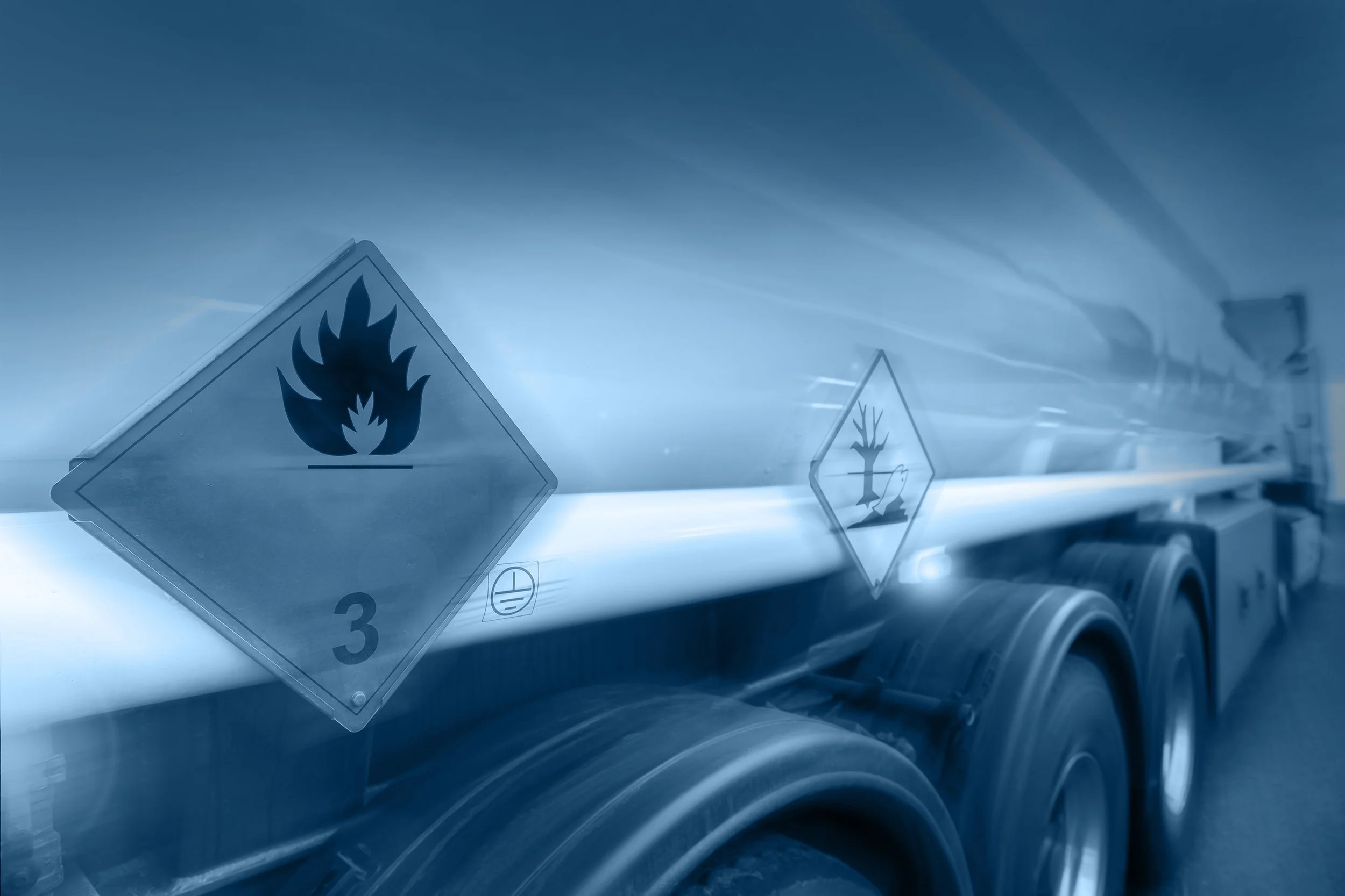 Close-up of a metal container with chemical hazard labels, including a flame symbol and a number 3 indicating flammable liquids, with the side of a vehicle or equipment in the background.