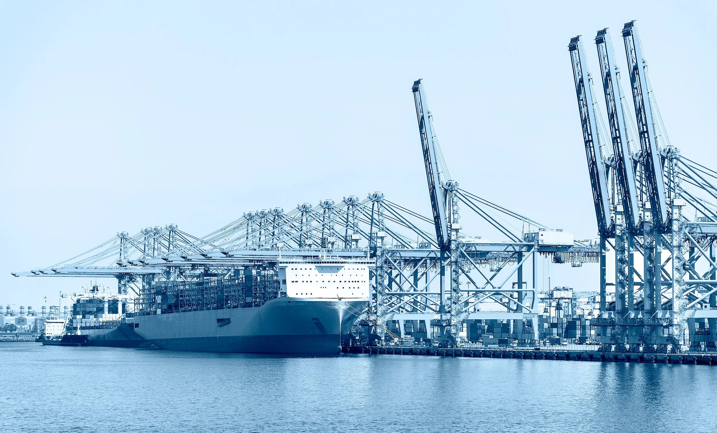 Cargo ships docked at a port with large cranes for loading and unloading containers, with water in the foreground and a clear sky.