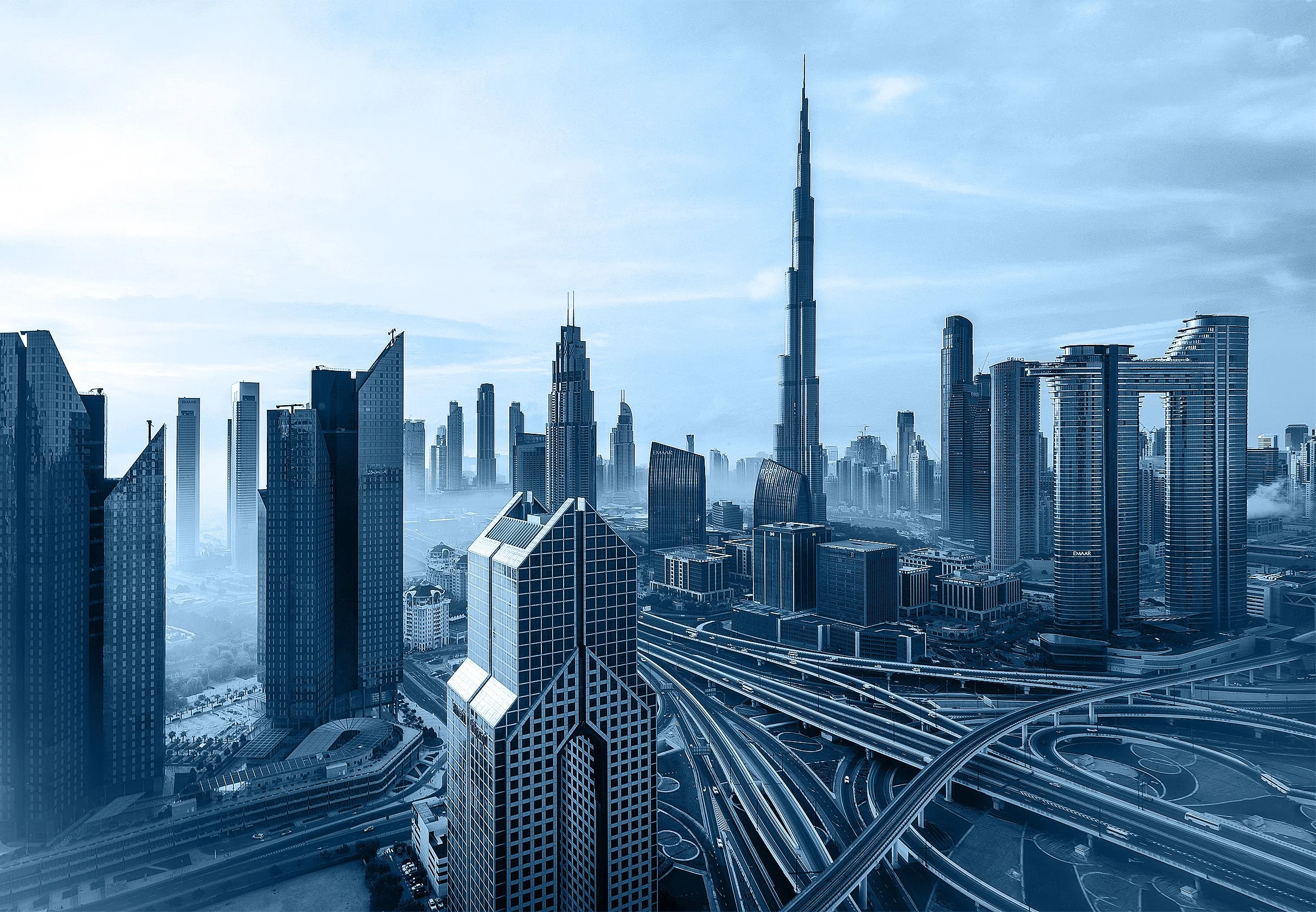 Dubai featuring numerous modern skyscrapers including the tall Burj Khalifa, under a partly cloudy sky. The image shows detailed road infrastructure with multiple highways weaving through the buildings.