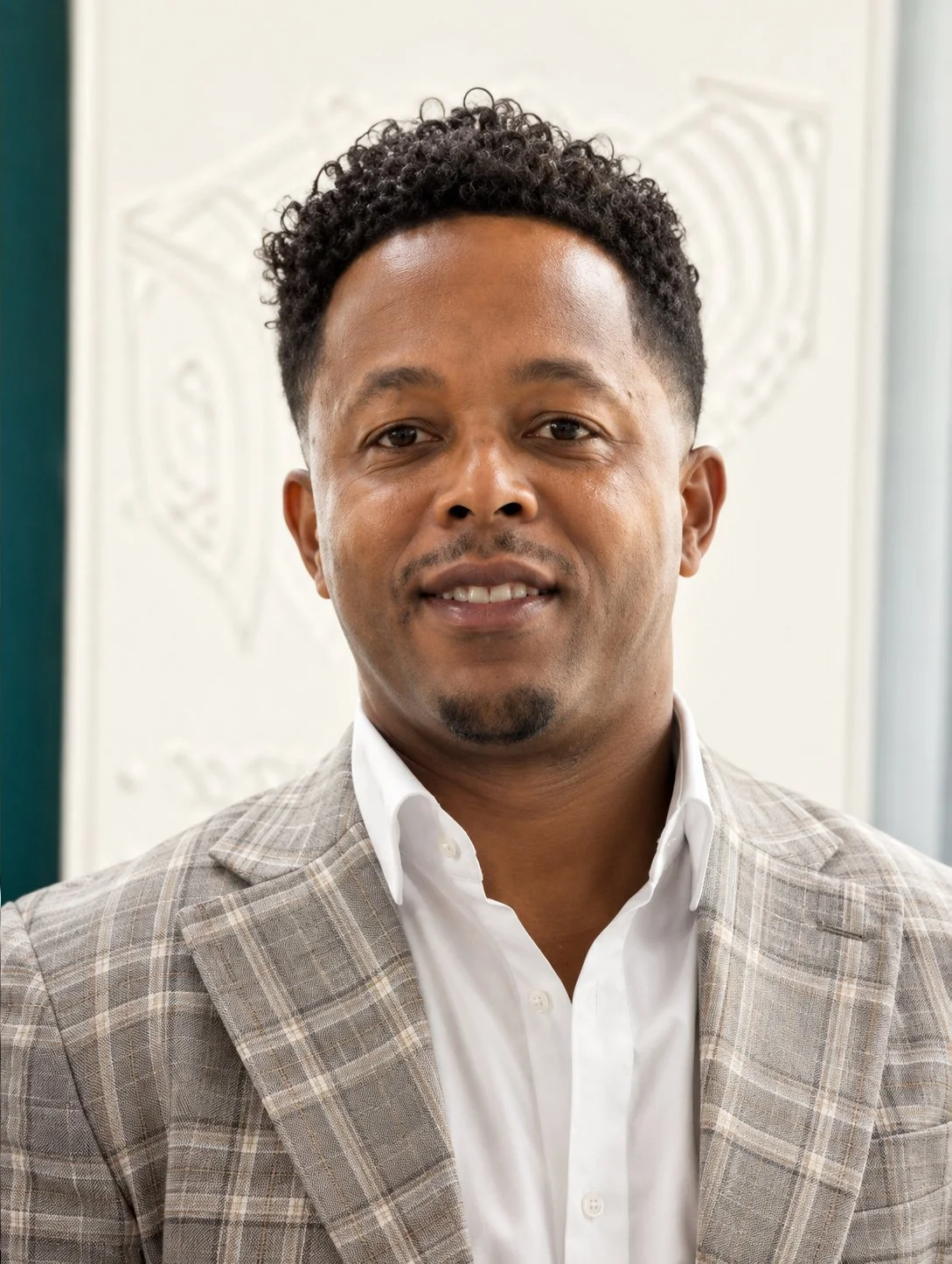 A man with curly black hair, wearing a beige checked blazer and white shirt, smiling at the camera.
