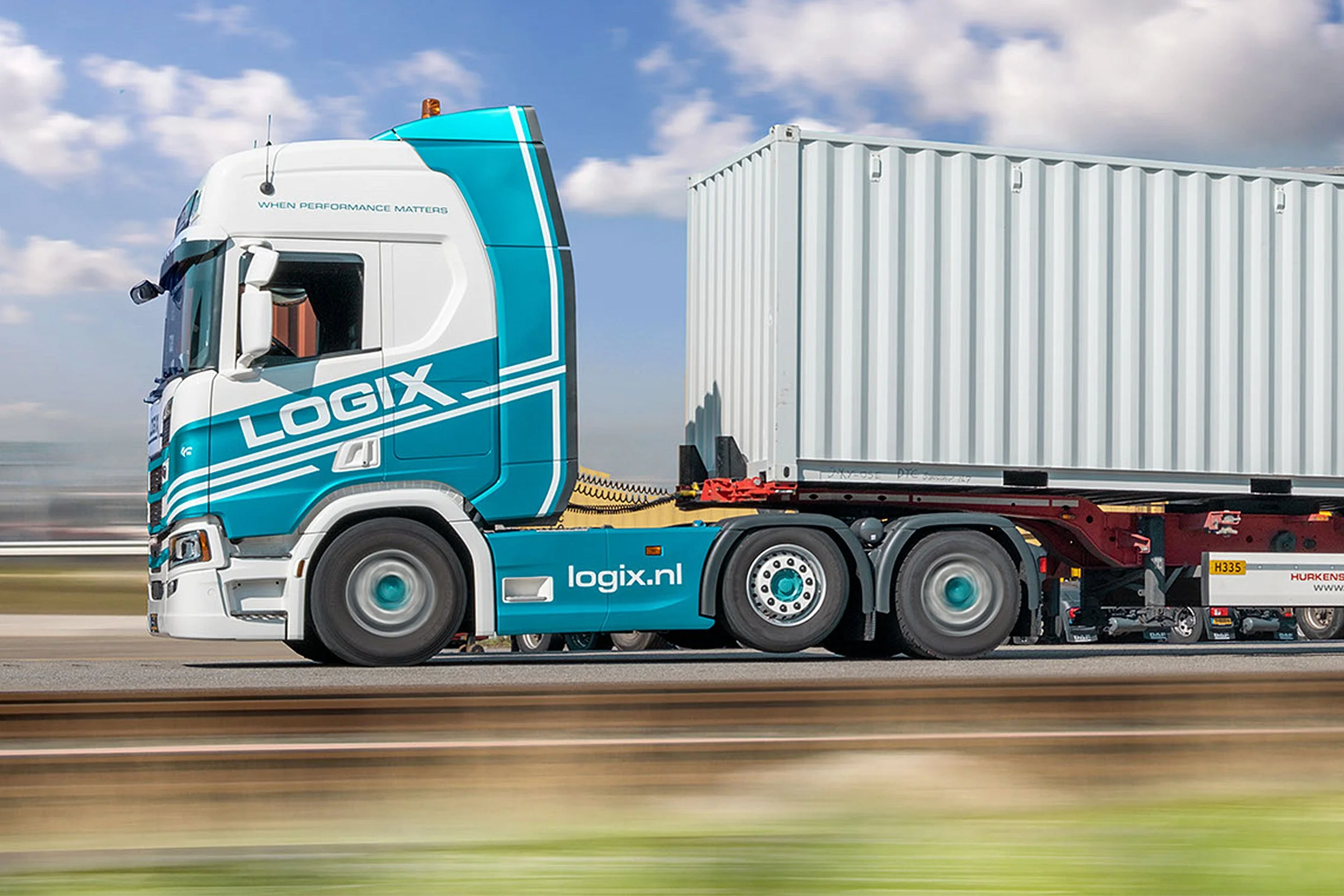 A blue and white Logix semi-truck hauling a large white shipping container on an open road with a partly cloudy sky in the background.