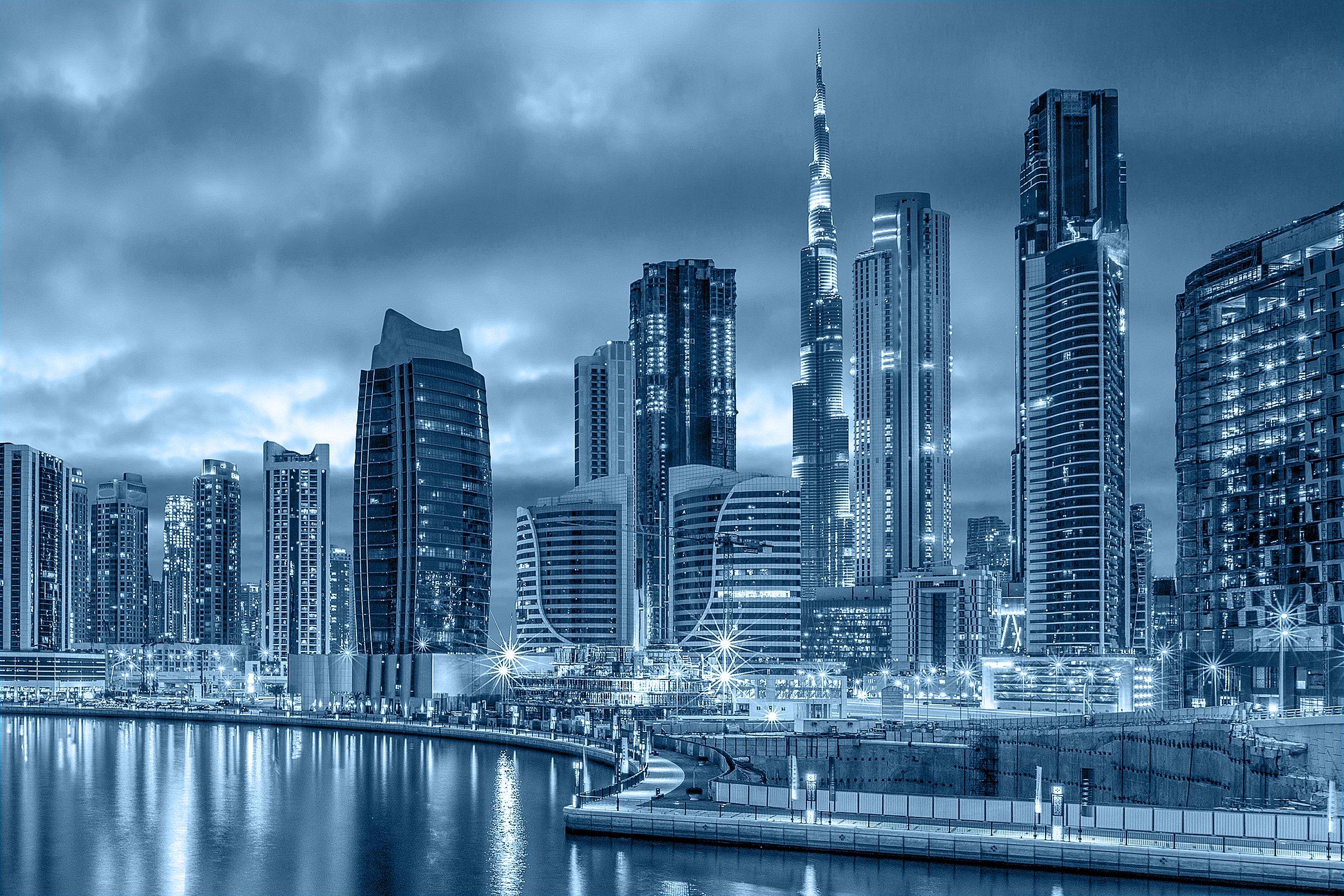 A city skyline with tall modern skyscrapers illuminated at night, reflecting on a body of water in the foreground, under a cloudy sky.