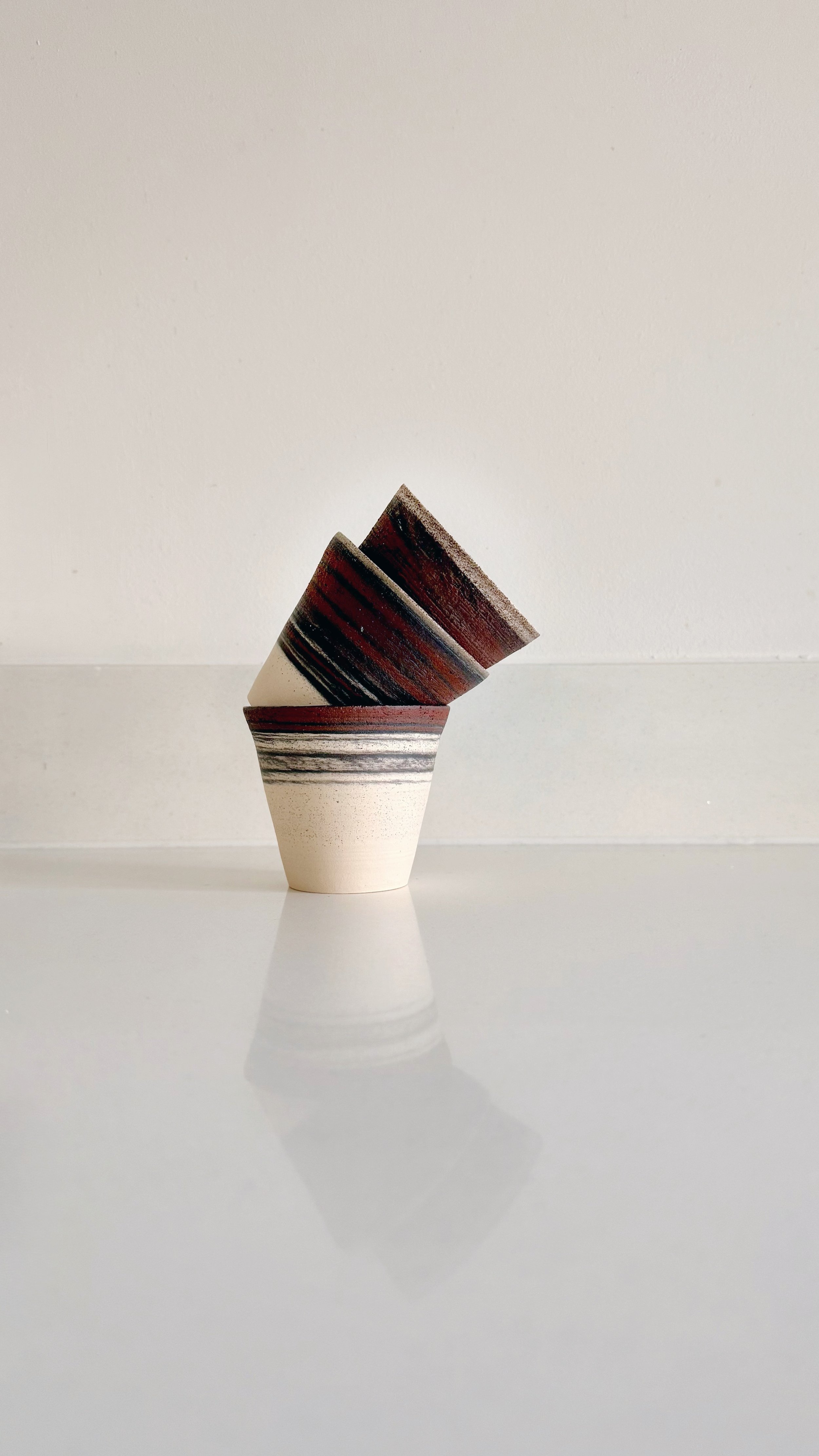 Three ceramic cups with black and red stripes, stacked inside each other, on a reflective surface against a plain white background.
