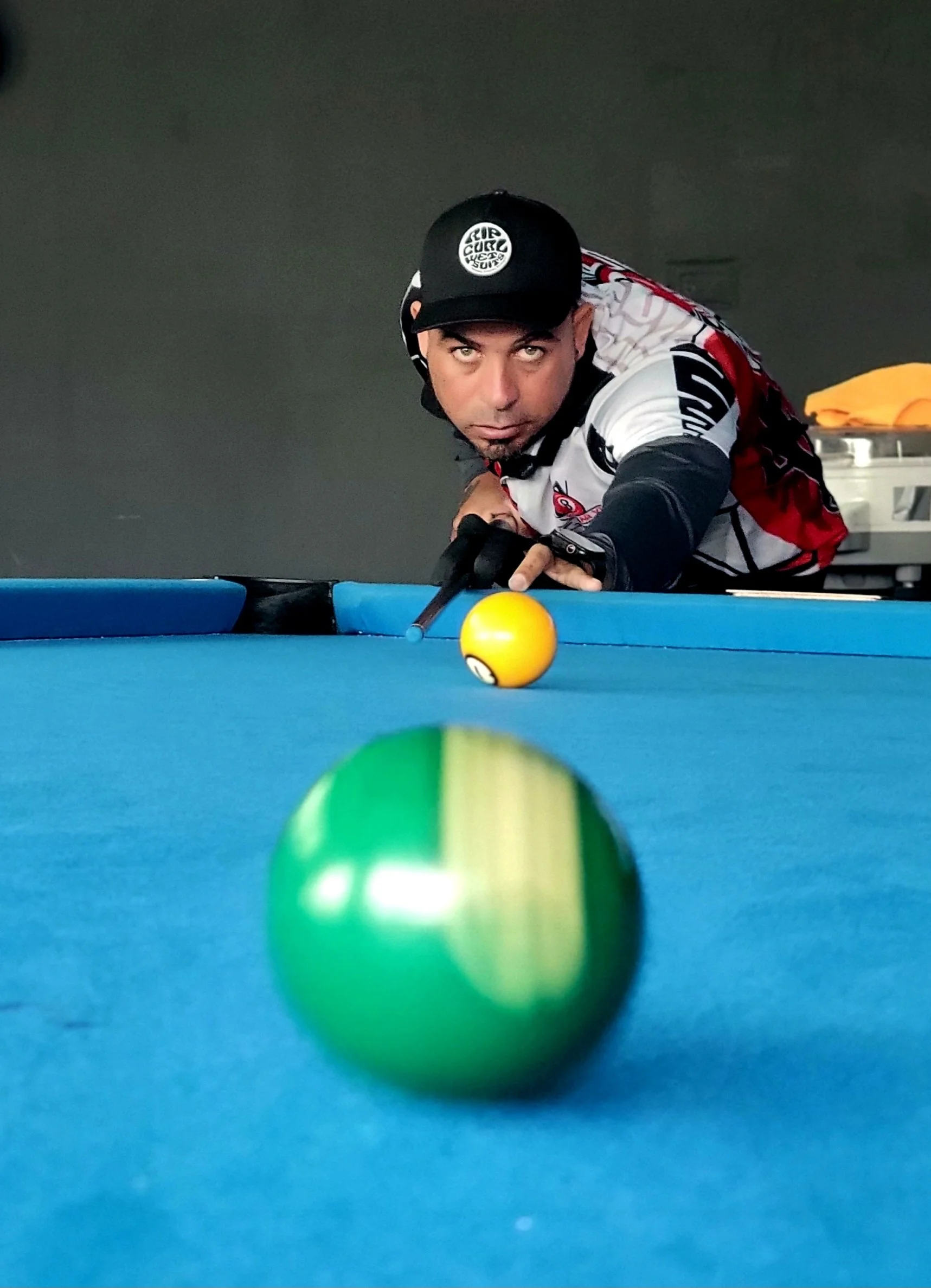 A man playing pool, aiming at the cue ball on a blue table, wearing a black cap, black gloves, and a sports jersey, with colorful balls in the foreground.
