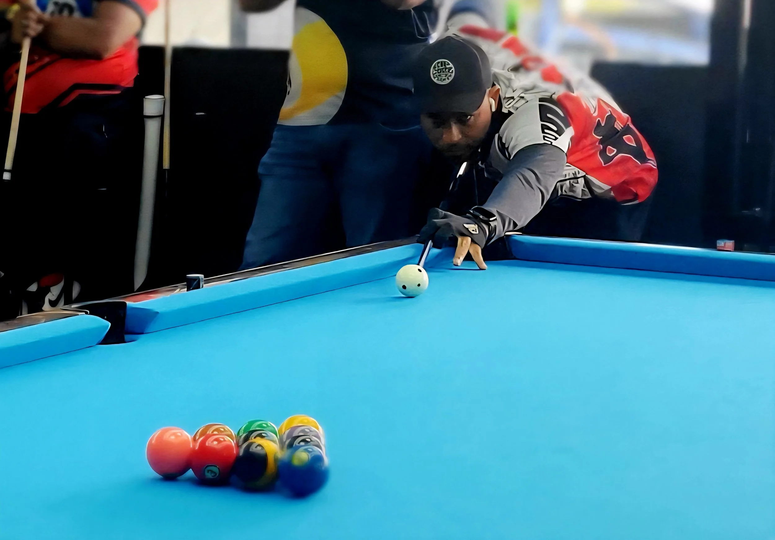 A man in a black cap and grey and red jacket leans over a blue billiards table, lining up to take a shot with a cue stick, with a white cue ball nearby and colorful billiard balls racked at the end of the table.