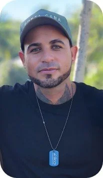 Close-up of a man wearing a baseball cap and a black shirt, outdoors with trees in the background.