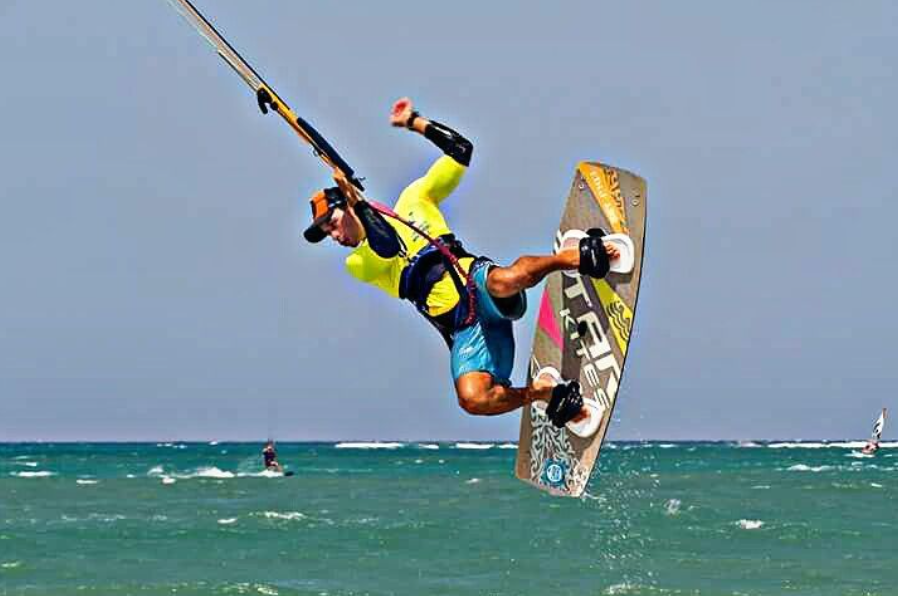 Kiteboarder performing aerial trick over ocean with other kiteboarders in the distance.