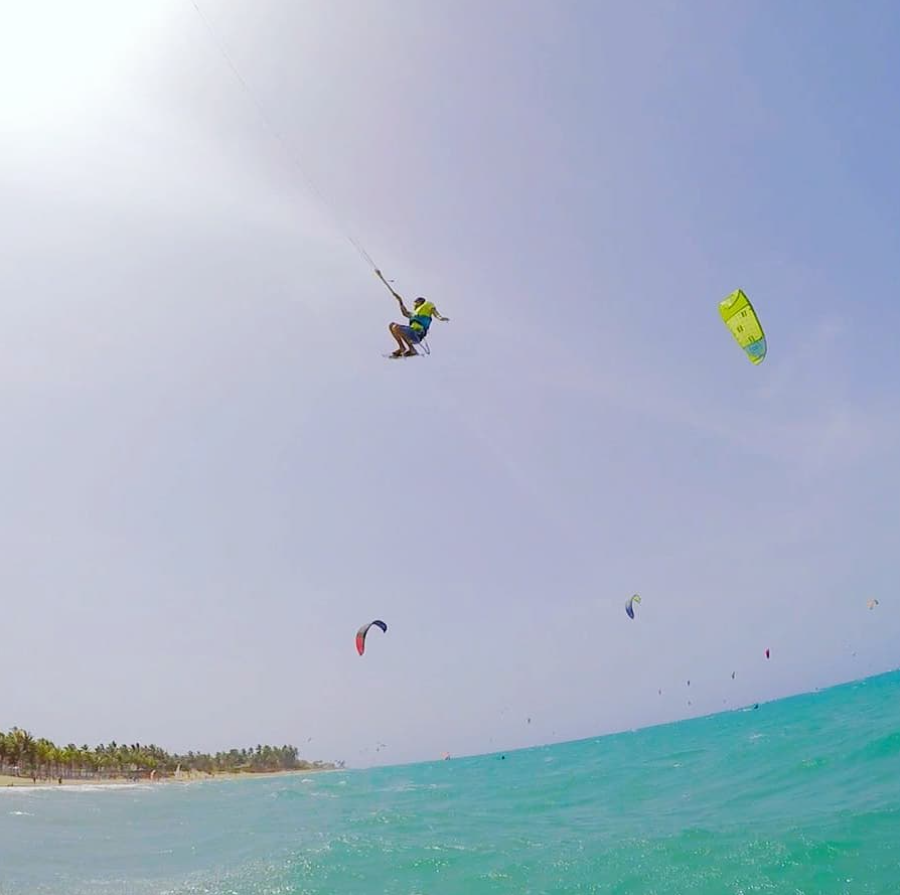 Person kiteboarding in the sky over the ocean with several colorful kite sails in the background and a distant shoreline with trees.