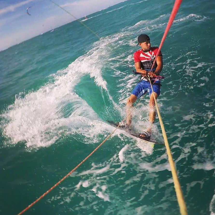 Young boy kiteboarding on the ocean, wearing a black cap, red and black shirt, blue shorts, and sandals, holding onto a kite control bar, with waves and distant kite surfers in the background.