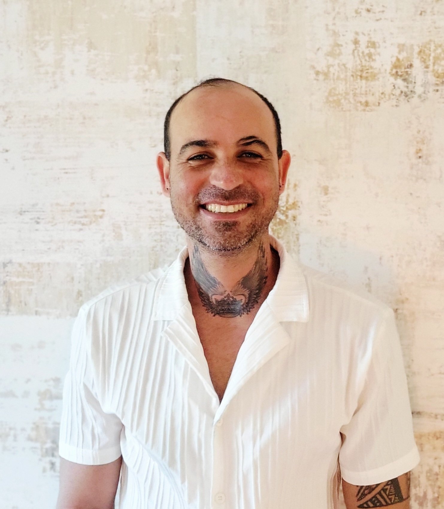 A smiling man with a shaved head, tattoos on his neck and arm, wearing a white, short-sleeved, collared shirt, standing in front of a textured beige wall.