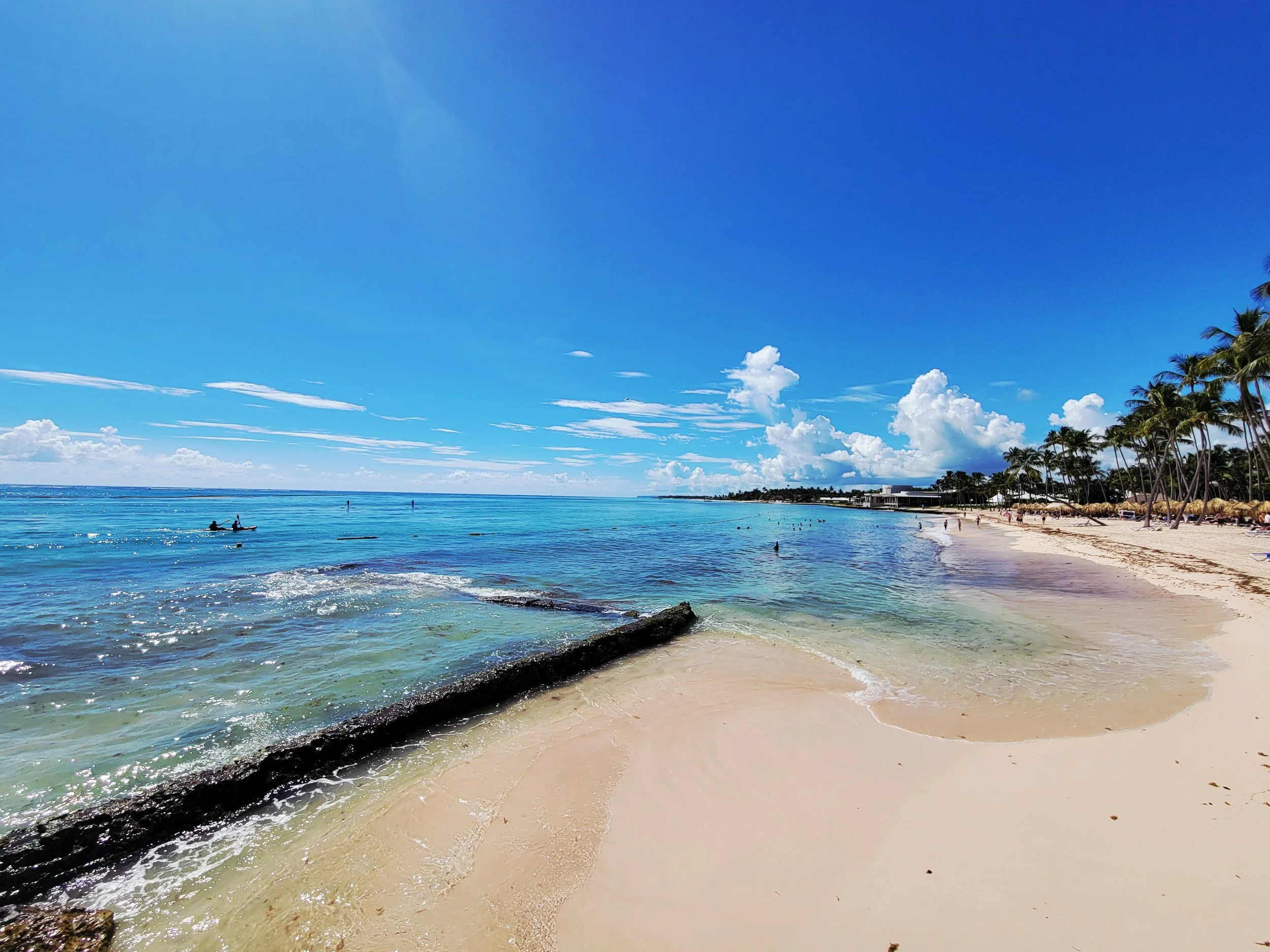 A sunny beach with turquoise water, palm trees lining the shore, and a few people swimming and kayaking in the ocean.