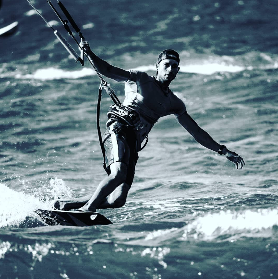 Man kiteboarding on the ocean, catching a wave, wearing a cap and sportswear.