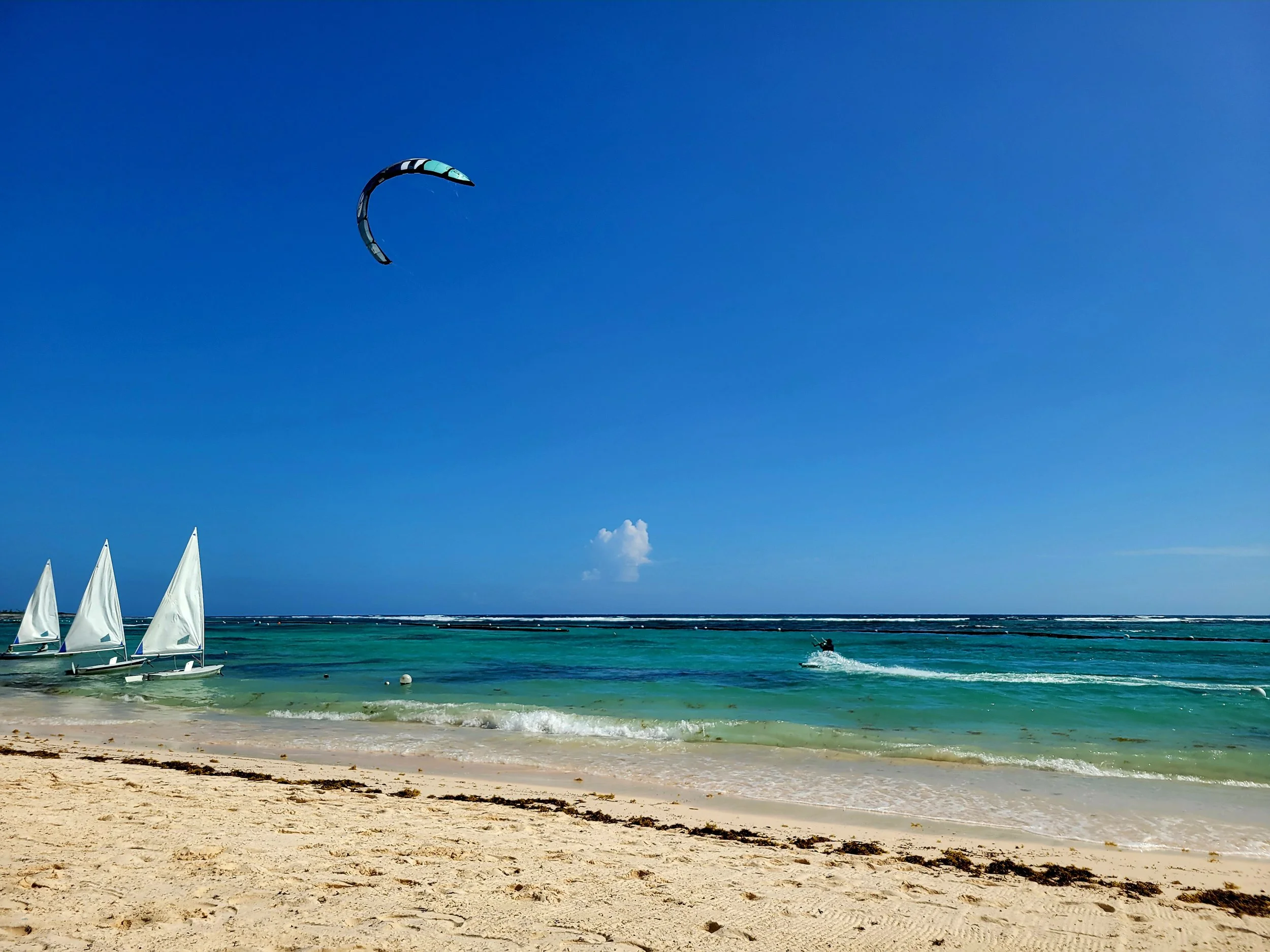 A beach with sailboats docked at the shore, a kite in the sky, and a person riding a jet ski on the water under a blue sky.