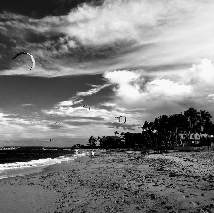 A black and white photo of a beach with four kite surfers flying their kites in the sky. Two people walk along the shoreline, with houses and palm trees visible in the background under a partly cloudy sky.