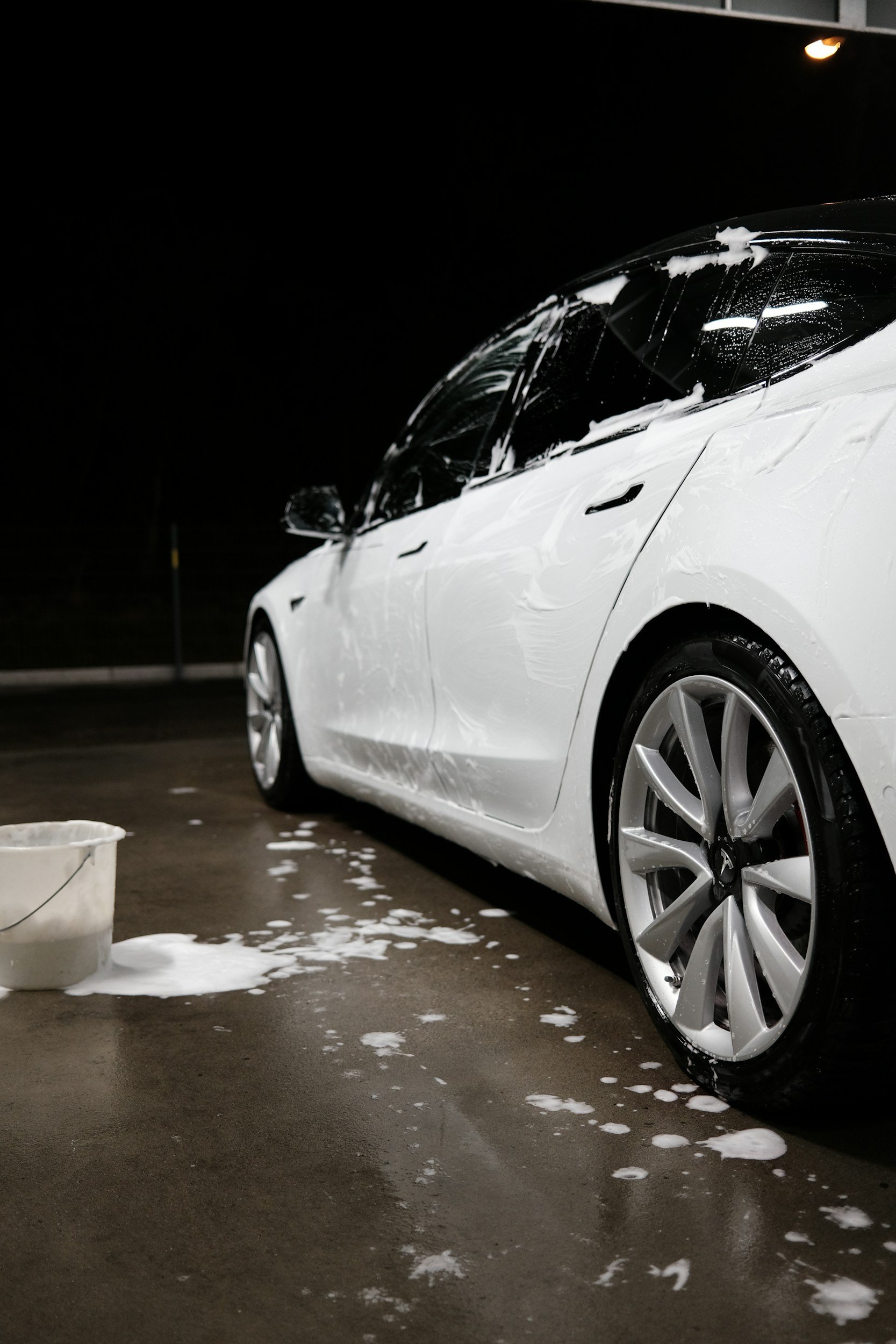 A white Tesla car is being washed outside at night, with foam and soap on the windshield and side. A bucket with soap suds is on the ground nearby, and soap bubbles and foam are spread across the wet pavement.