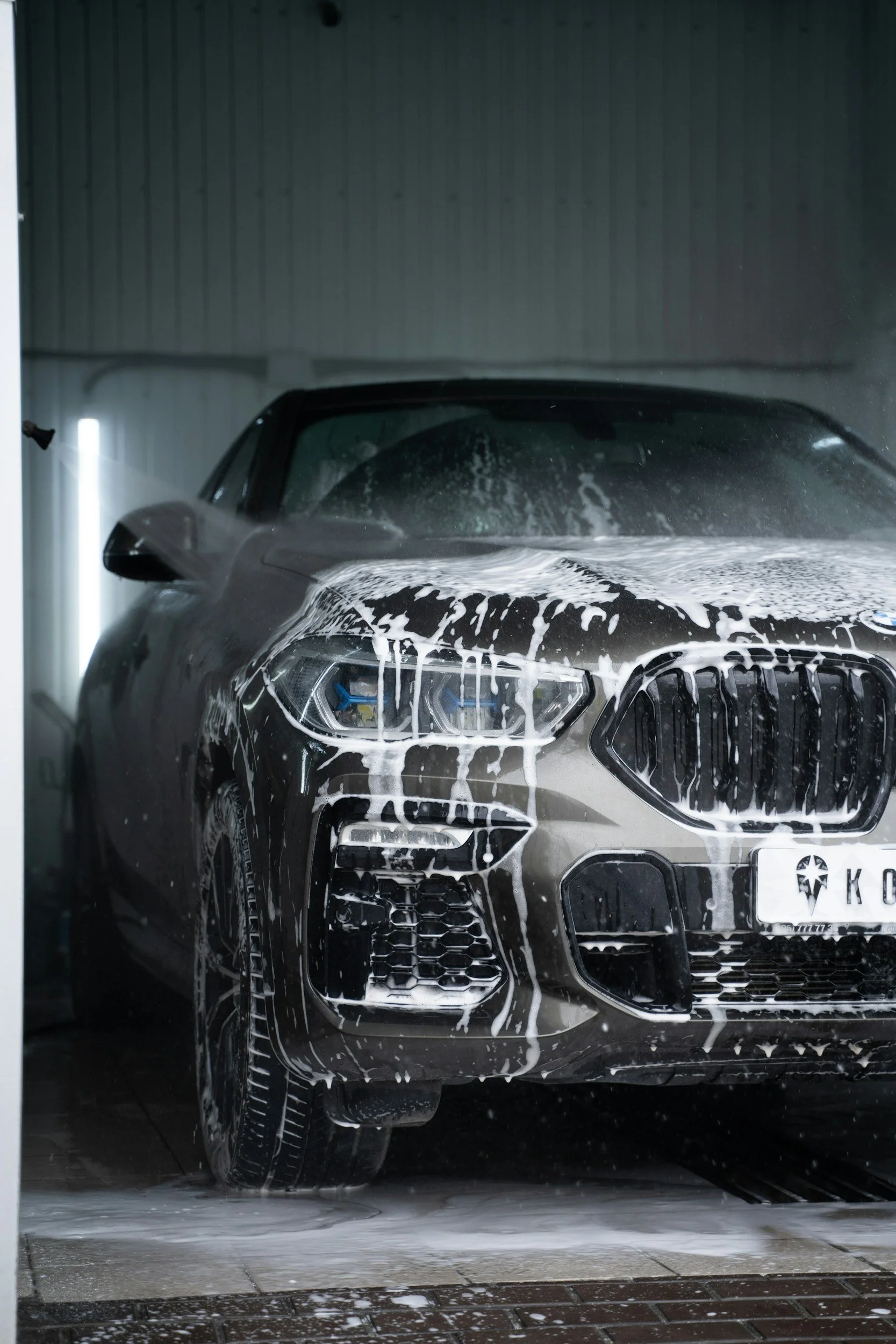 Black luxury car in a car wash with soap and water foam on the front, headlights, and grille, inside a garage with tiled floor.