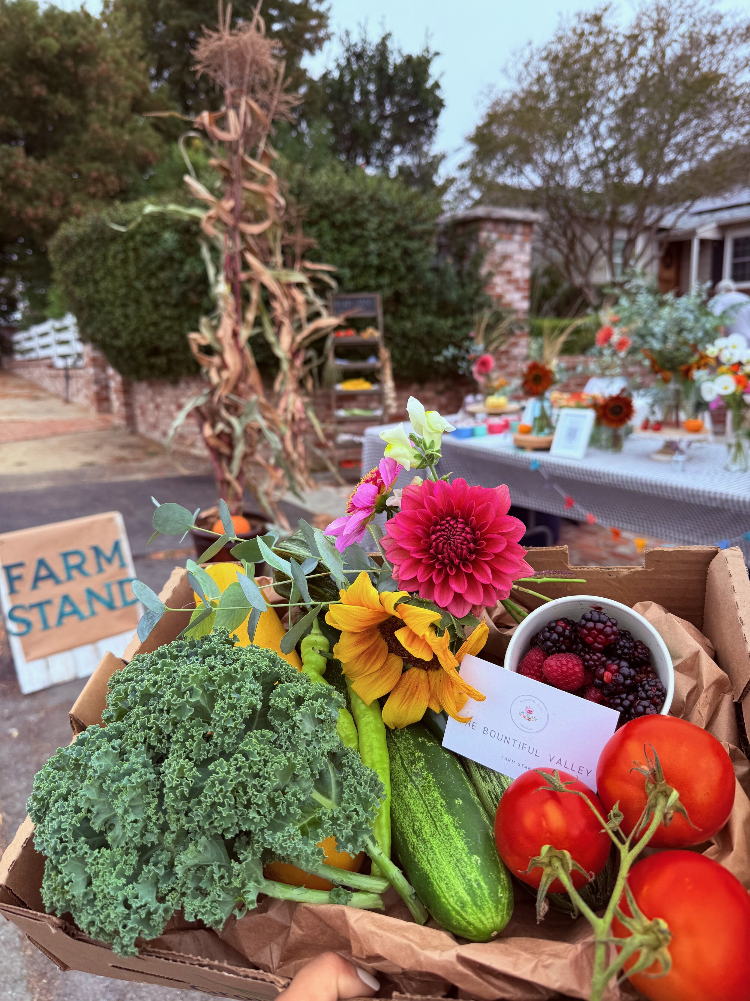 A box of fresh produce including tomatoes, cucumbers, lettuce, and a cup of mixed berries, with colorful flowers, at a farm stand.