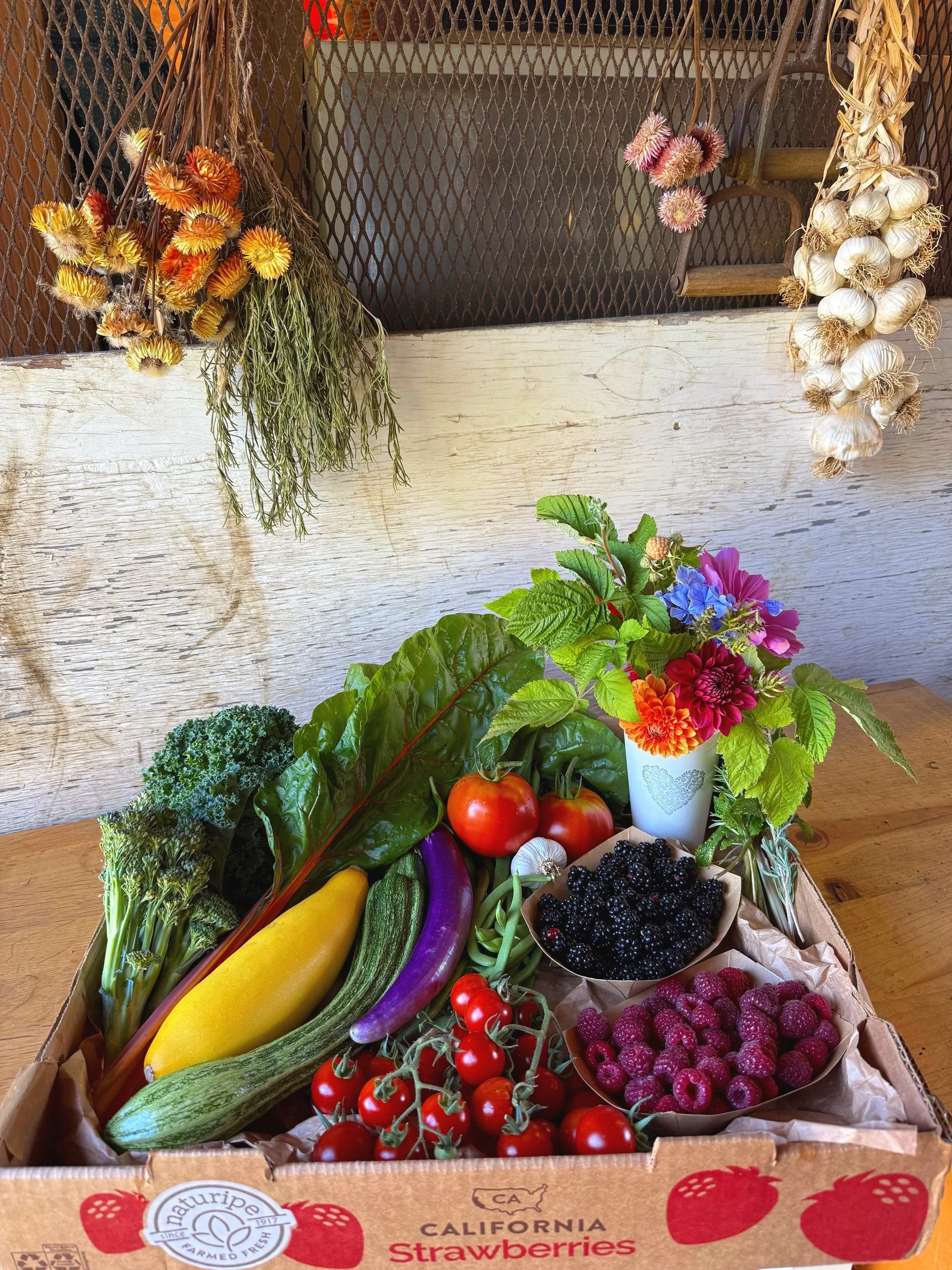 A cardboard box filled with fresh vegetables and berries, including tomatoes, eggplant, zucchini, broccoli, and berries, on a wooden table, with colorful flowers in a white mug.