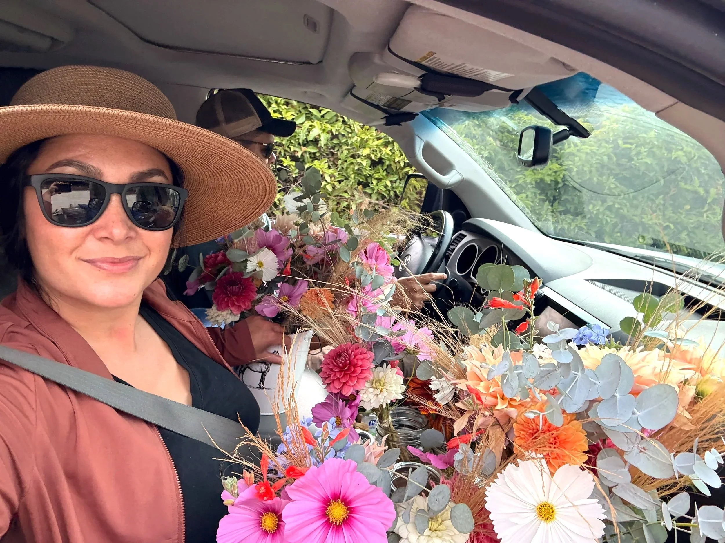 Woman wearing brown jacket, black shirt, sunglasses, and wide-brimmed hat, sitting in a vehicle filled with colorful flowers and greenery visible outside the window.