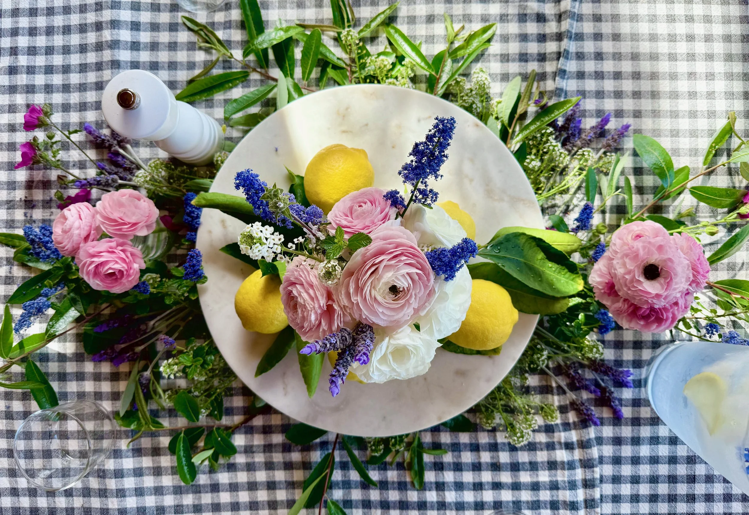 A floral centerpiece with pink and white flowers, blue lavender, and green leaves on a checkered tablecloth, with lemons and a white candle nearby.