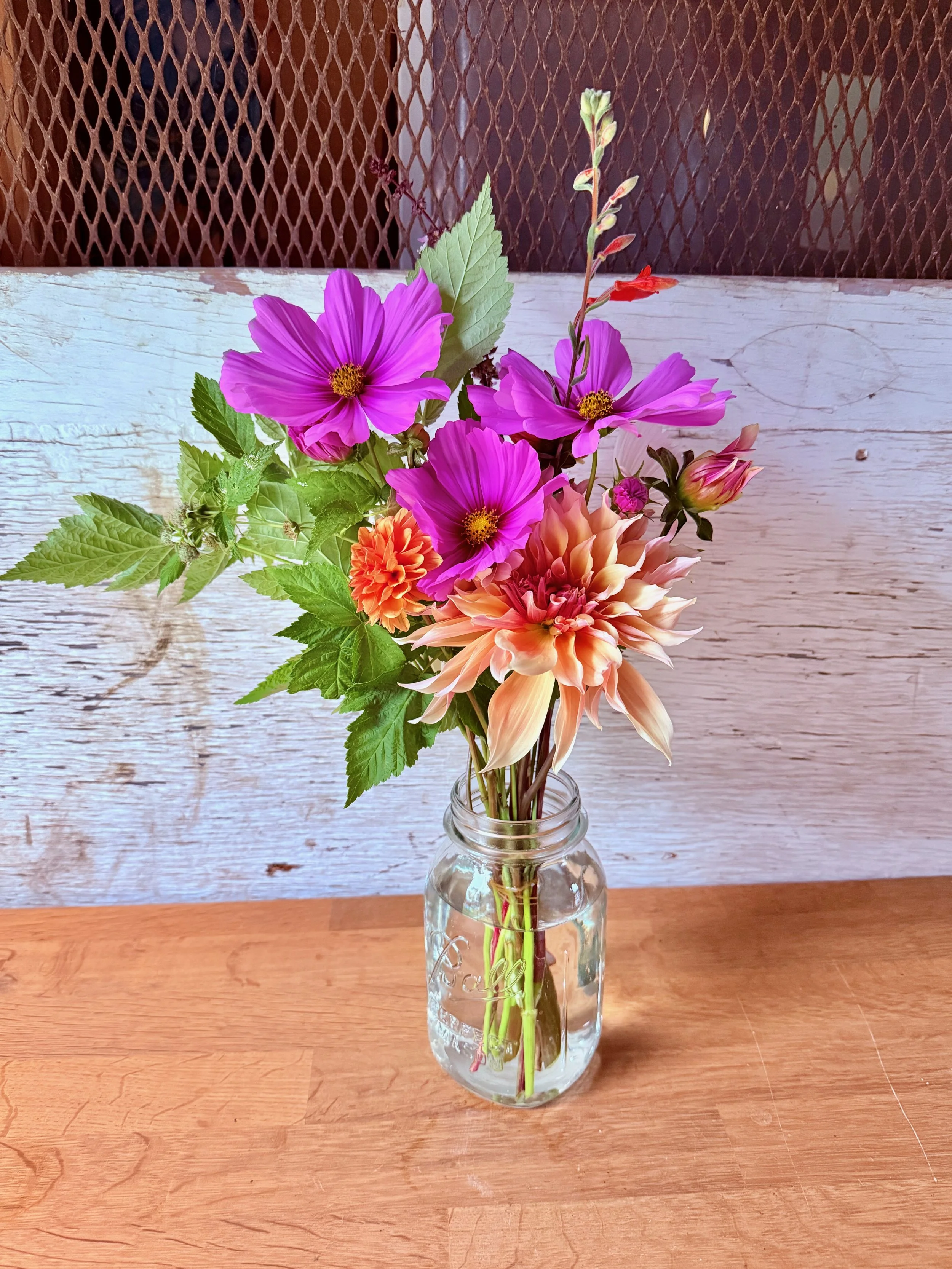 A glass vase filled with a colorful bouquet of pink, orange, and peach flowers, with green leaves, set on a wooden table against a rustic white wooden background.