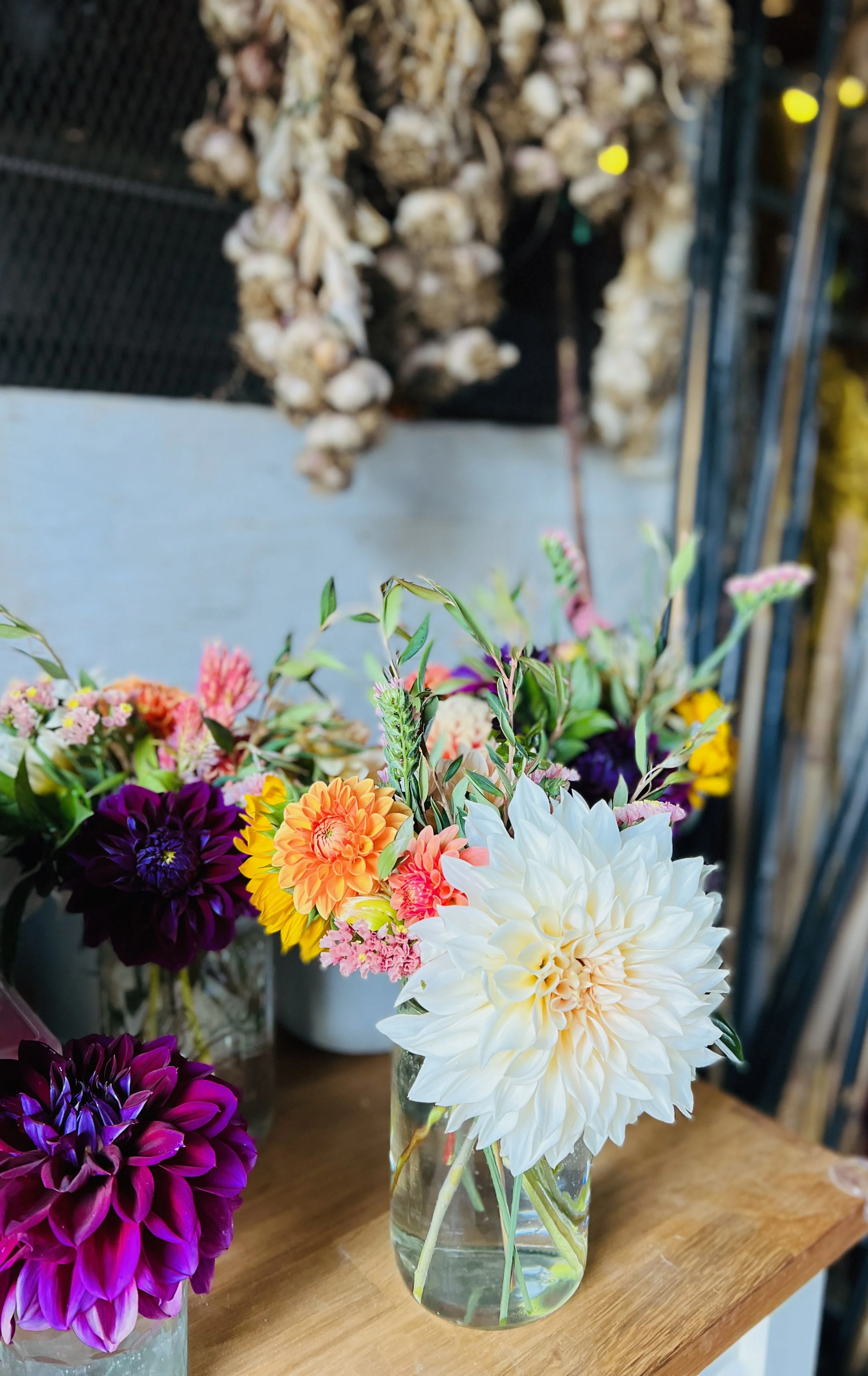 Vase of mixed fresh flowers including a large white, orange, pink, and purple dahlias, on a wooden surface, with garlic bunches hanging in the background.
