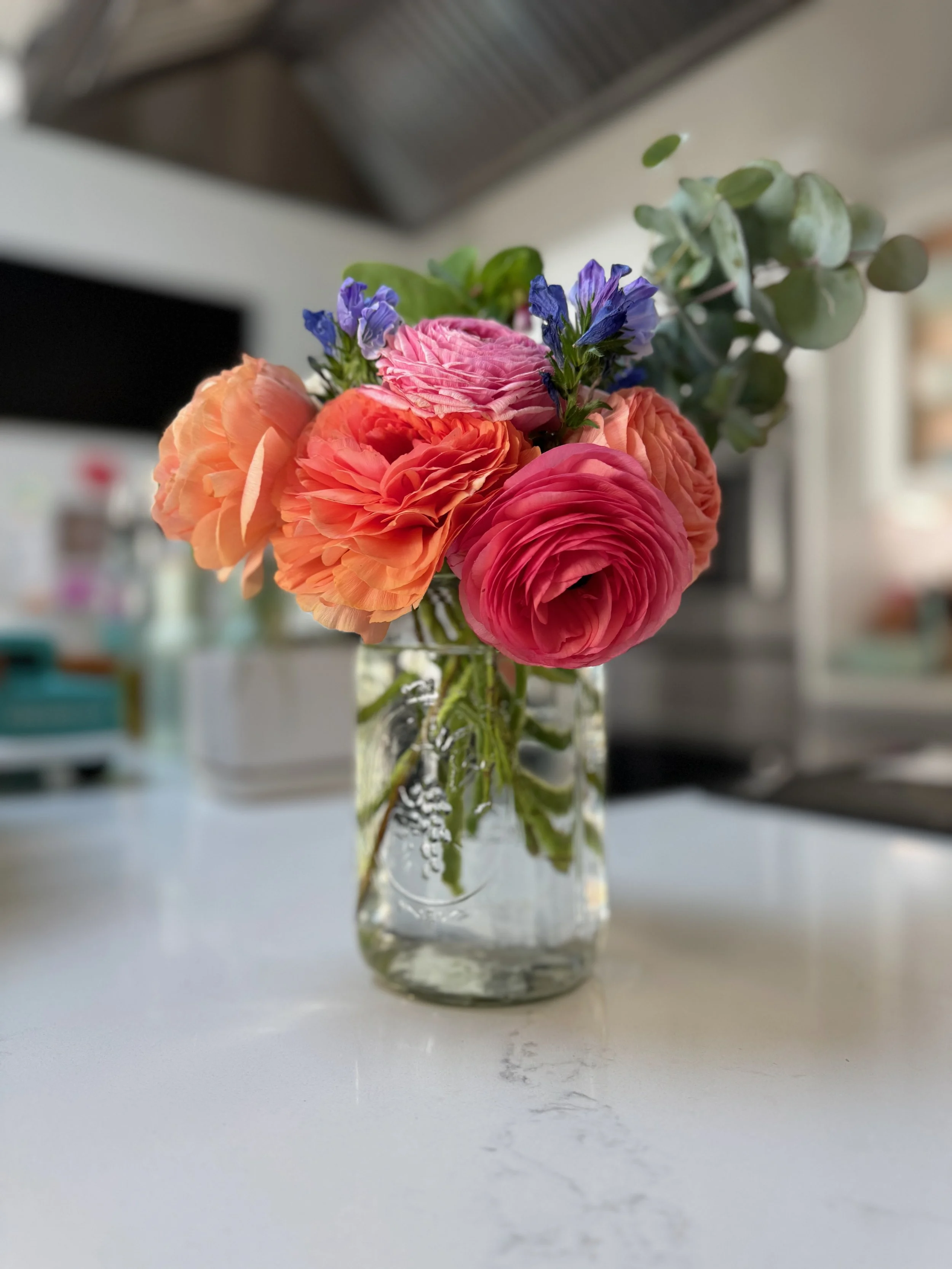 A glass vase with colorful flowers, including pink, orange, and purple blossoms, and some green foliage, on a white surface in a bright room.