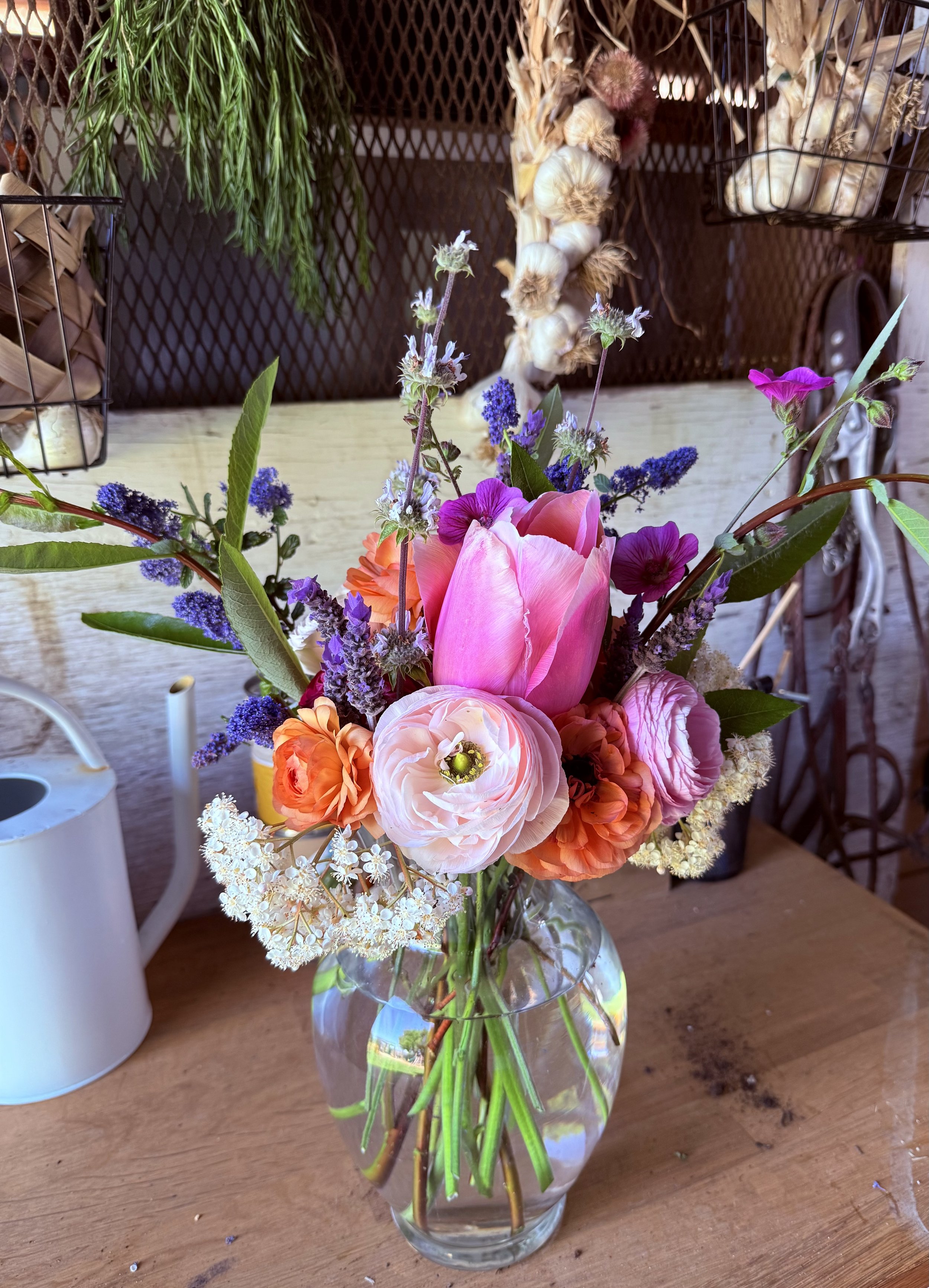 A colorful bouquet of various flowers including pink tulips, orange blooms, white and purple blossoms, arranged in a clear glass vase on a wooden table.