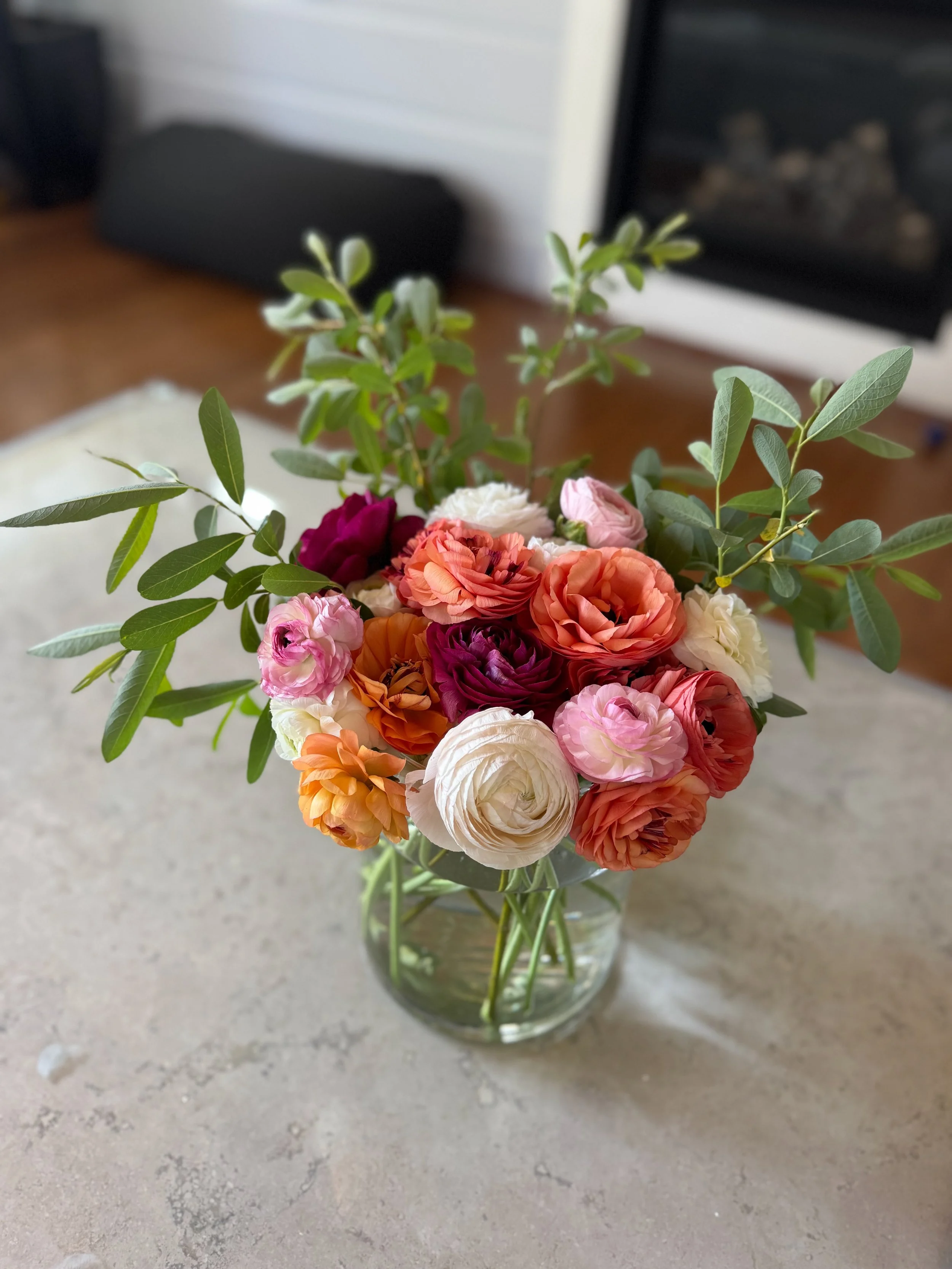 A glass vase filled with a variety of colorful flowers, including pink, white, orange, and burgundy blooms, and green leafy stems, placed on a beige surface in a home setting.