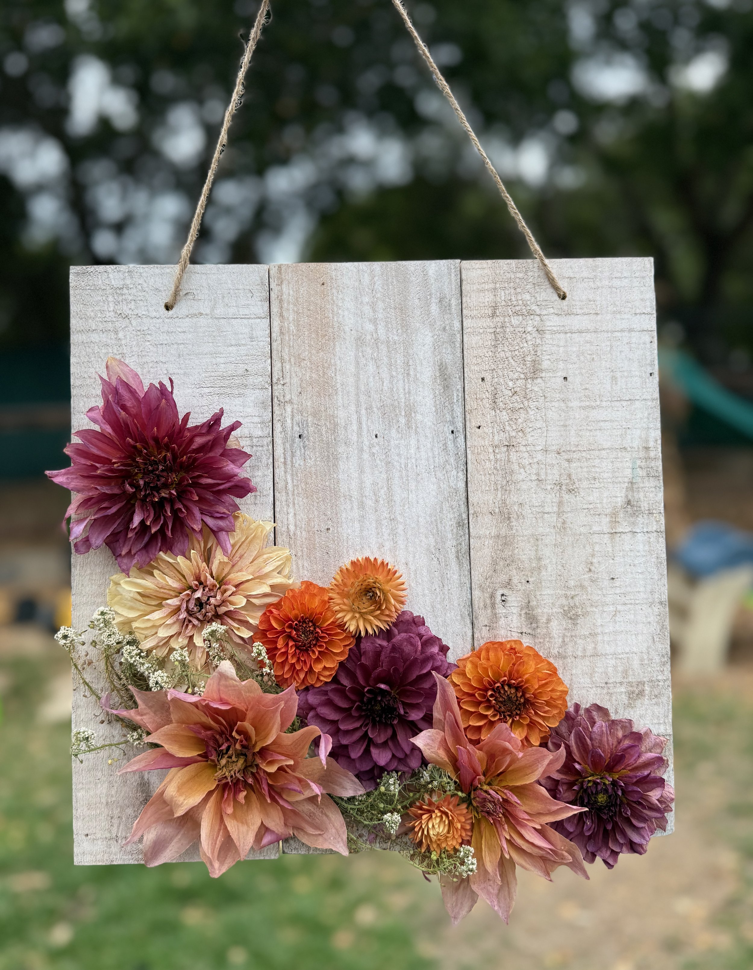 Hanging wooden sign decorated with pink, orange, peach, and purple flowers, outdoors with blurred green trees in the background.