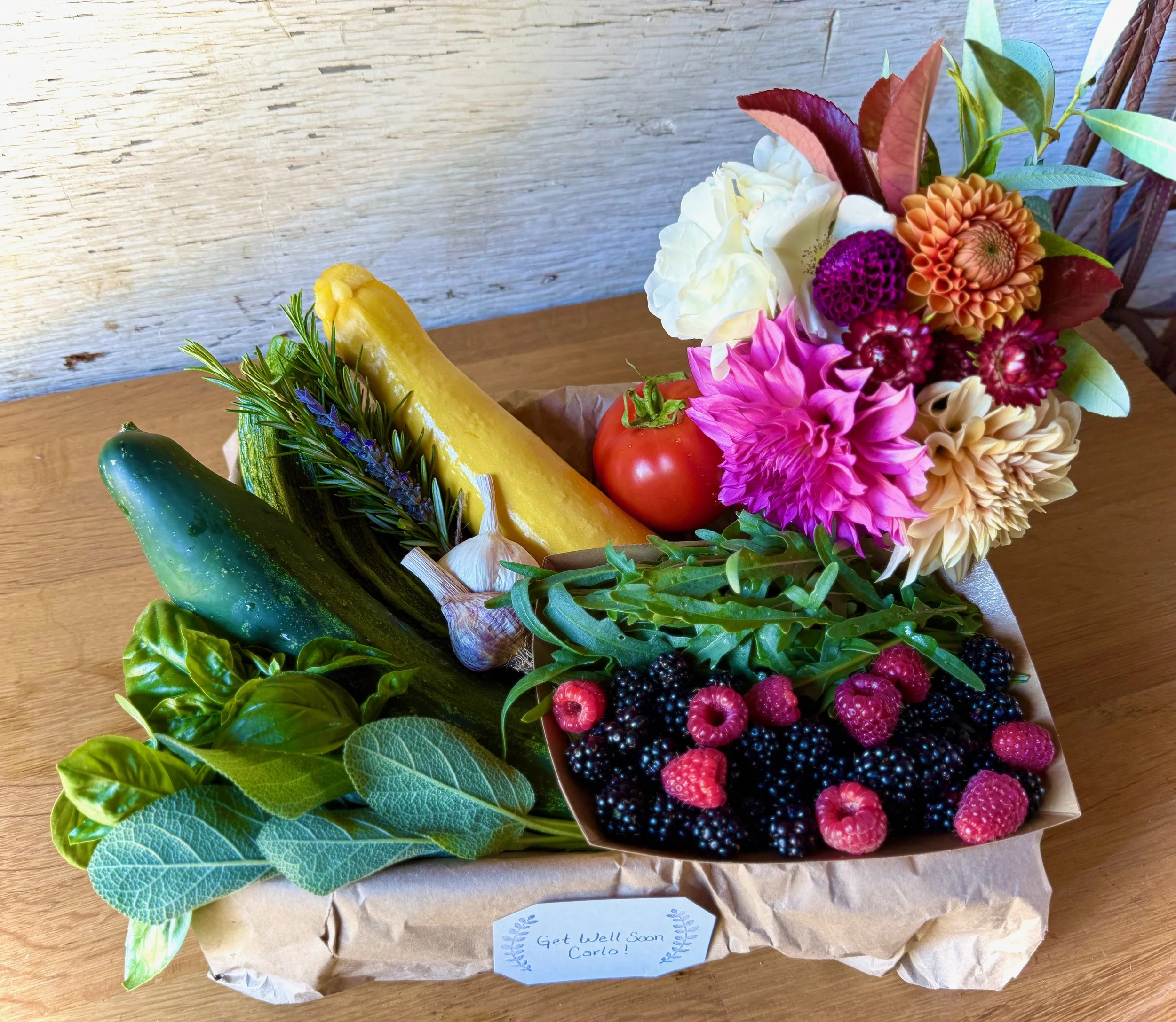 A gift basket with fresh vegetables, herbs, a tomato, mixed berries, and a colorful bouquet of flowers, with a personal note.
