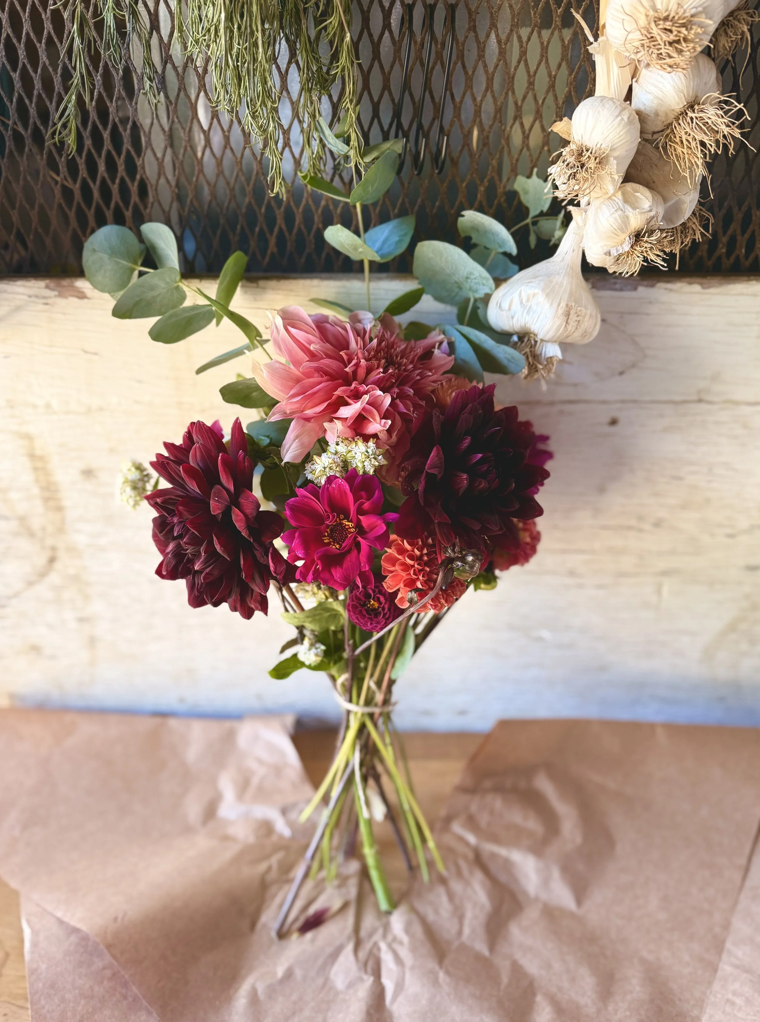 A bouquet of pink, deep red, and magenta flowers with green leaves and small white filler flowers, placed on brown paper on a wooden surface.