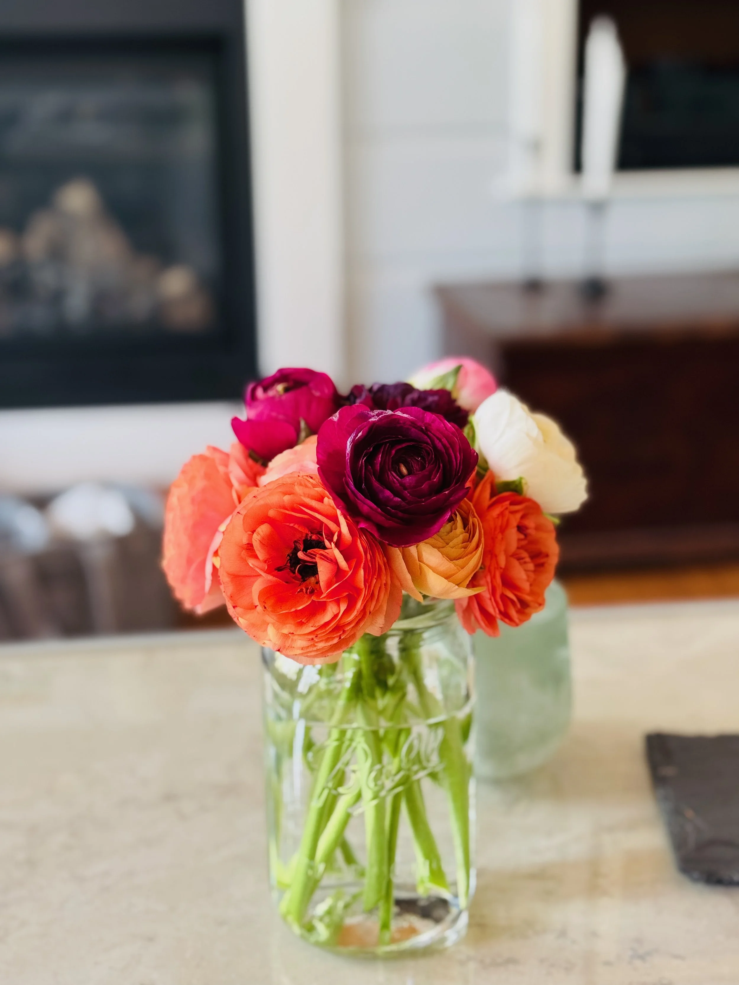 A bouquet of colorful ranunculus flowers in a glass vase on a beige surface.