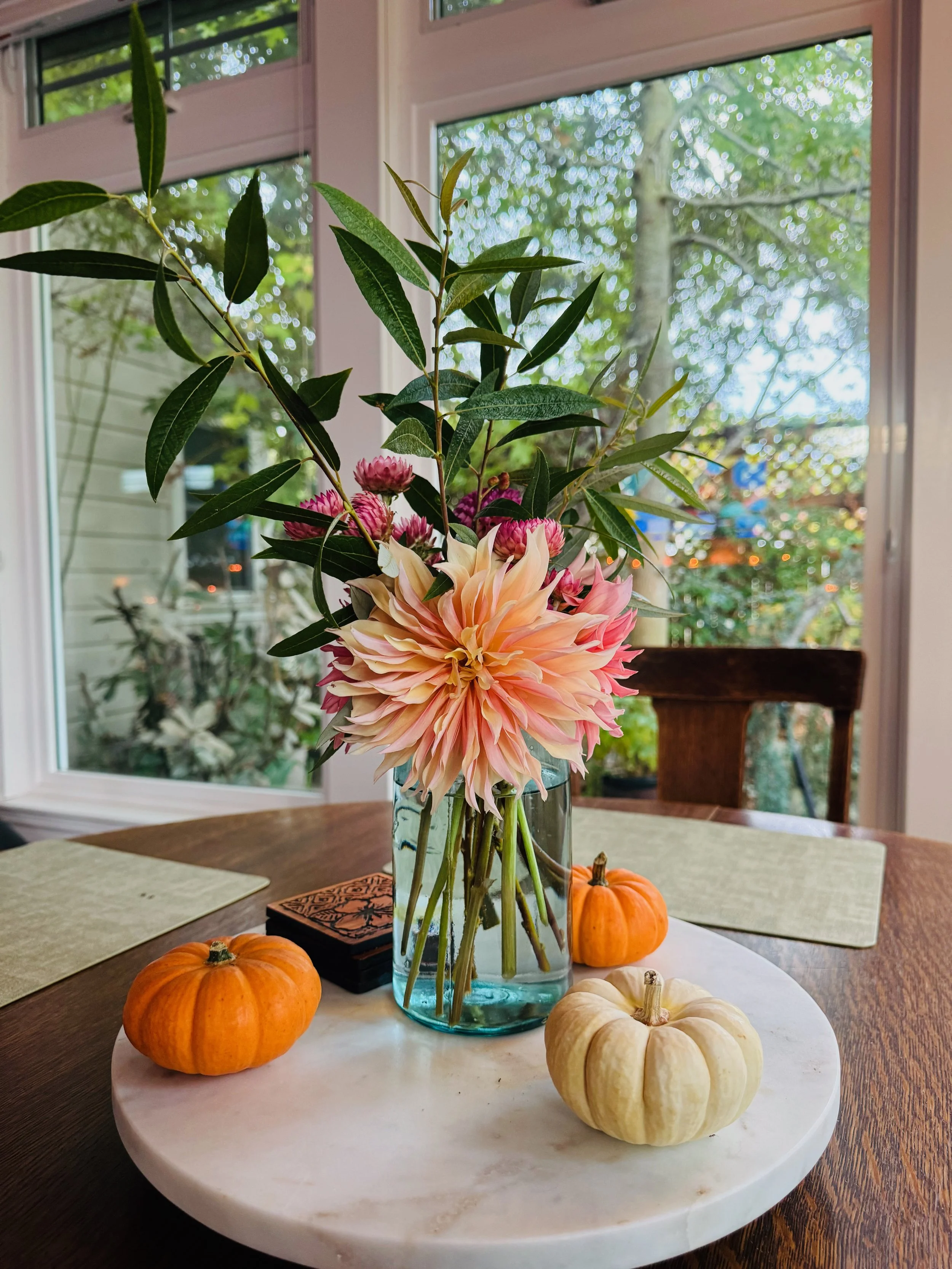 A floral arrangement in a glass vase on a white marble circular tray, with three small pumpkins around it on a wood dining table, in a sunlit room with large windows and green foliage outside.