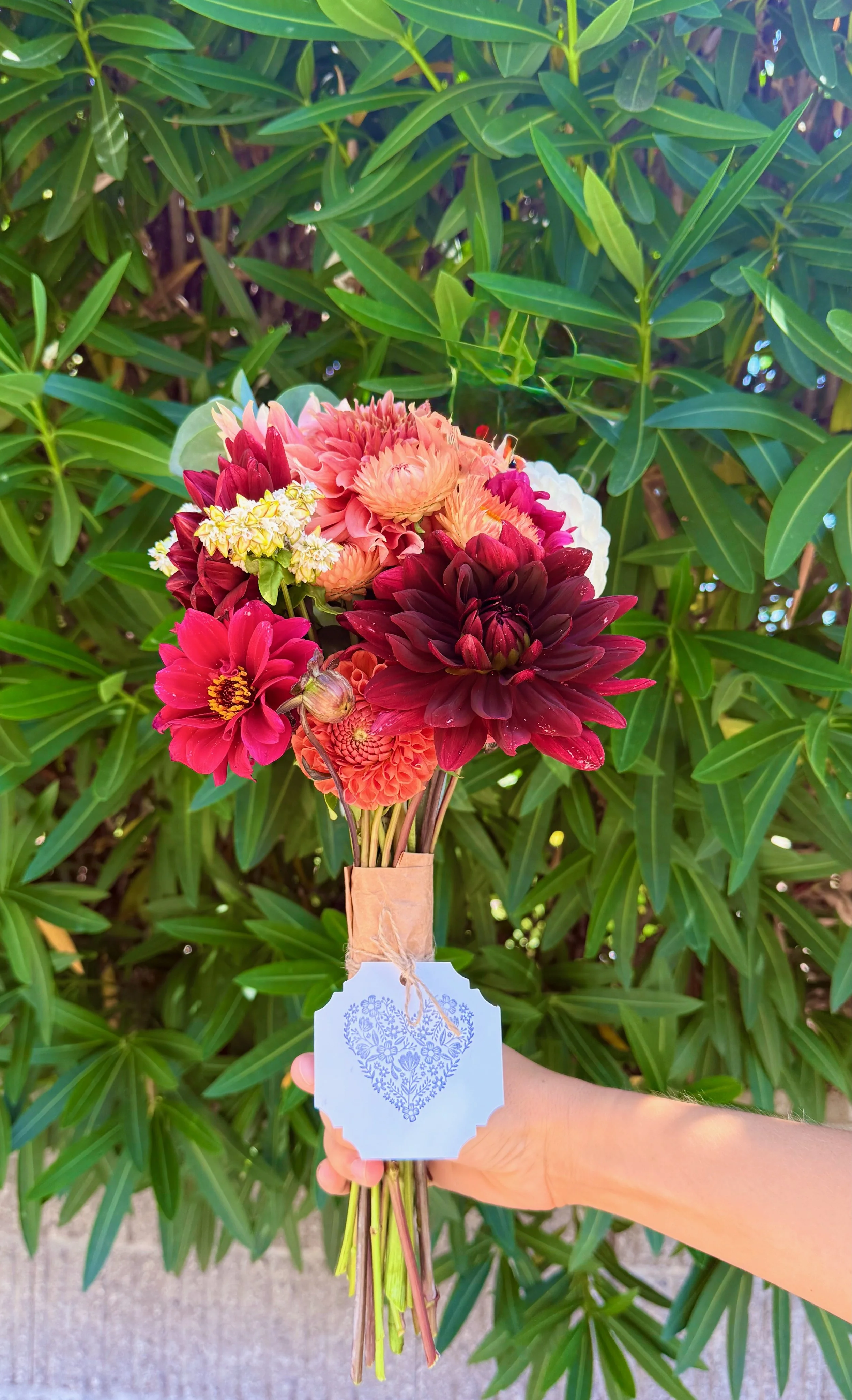 Hand holding a bouquet of pink, red, and peach flowers with a blue heart-shaped tag against a backdrop of green leaves.