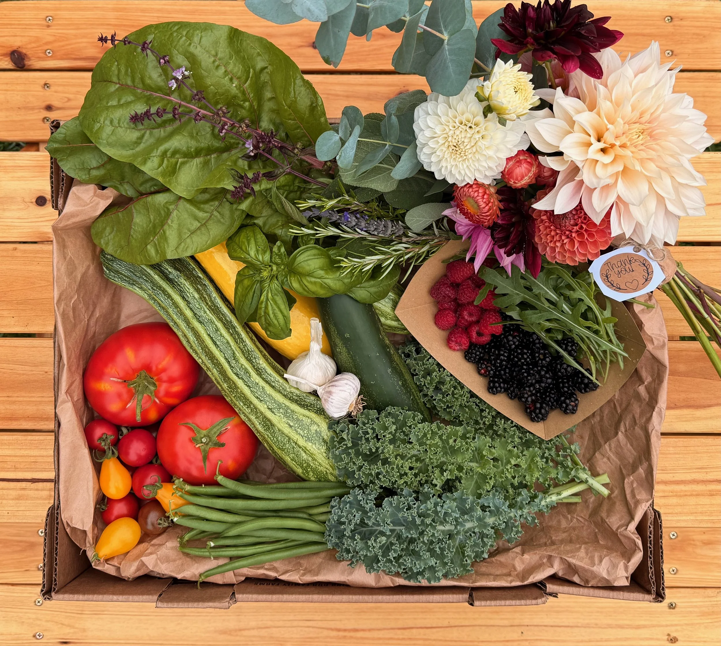 Fresh vegetables, herbs, and a bouquet of flowers arranged in a box on a wooden surface. The vegetables include tomatoes, zucchini, garlic, green beans, kale, and a yellow squash. The herbs include basil, thyme, mint, and lavender. The bouquet has da