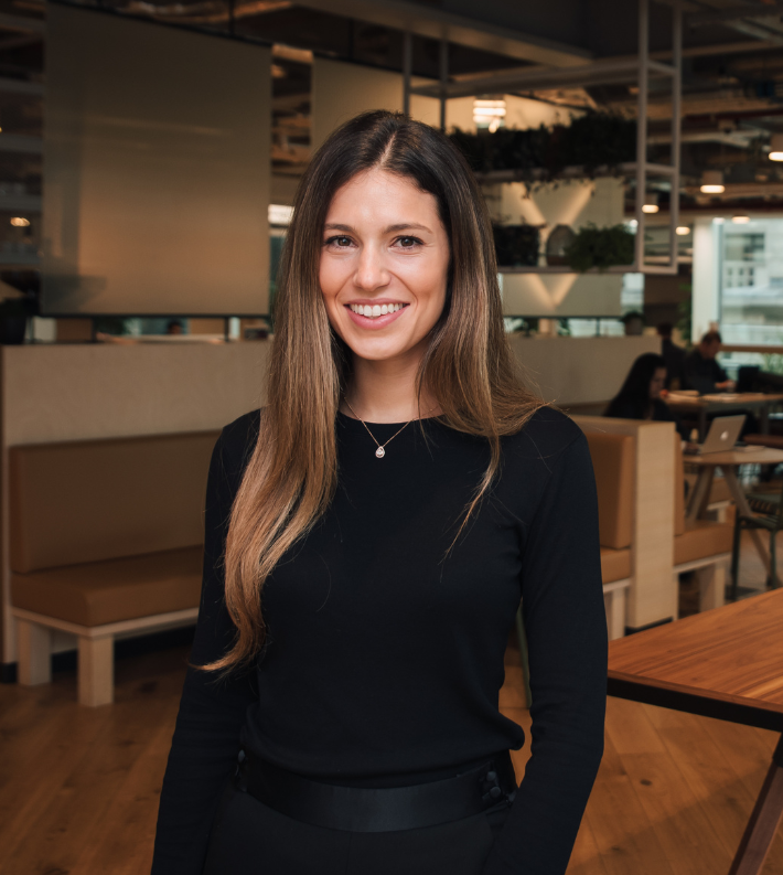 A young woman with long brown hair and a black long-sleeve top, smiling in a modern office cafe.