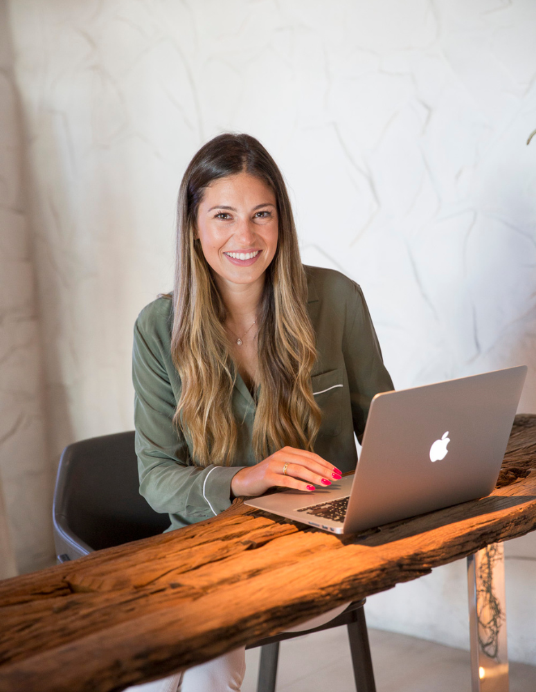 A woman with long, wavy hair sitting at a rustic wooden table, smiling at the camera with a silver MacBook laptop in front of her.