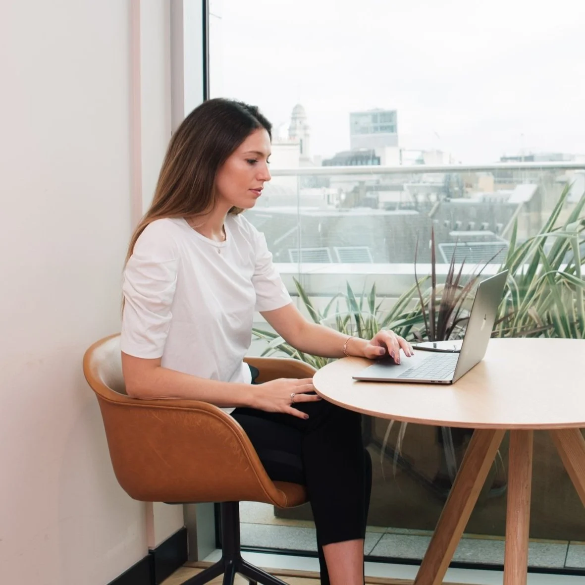 Woman working on a laptop at a round table near a large window with a city view.