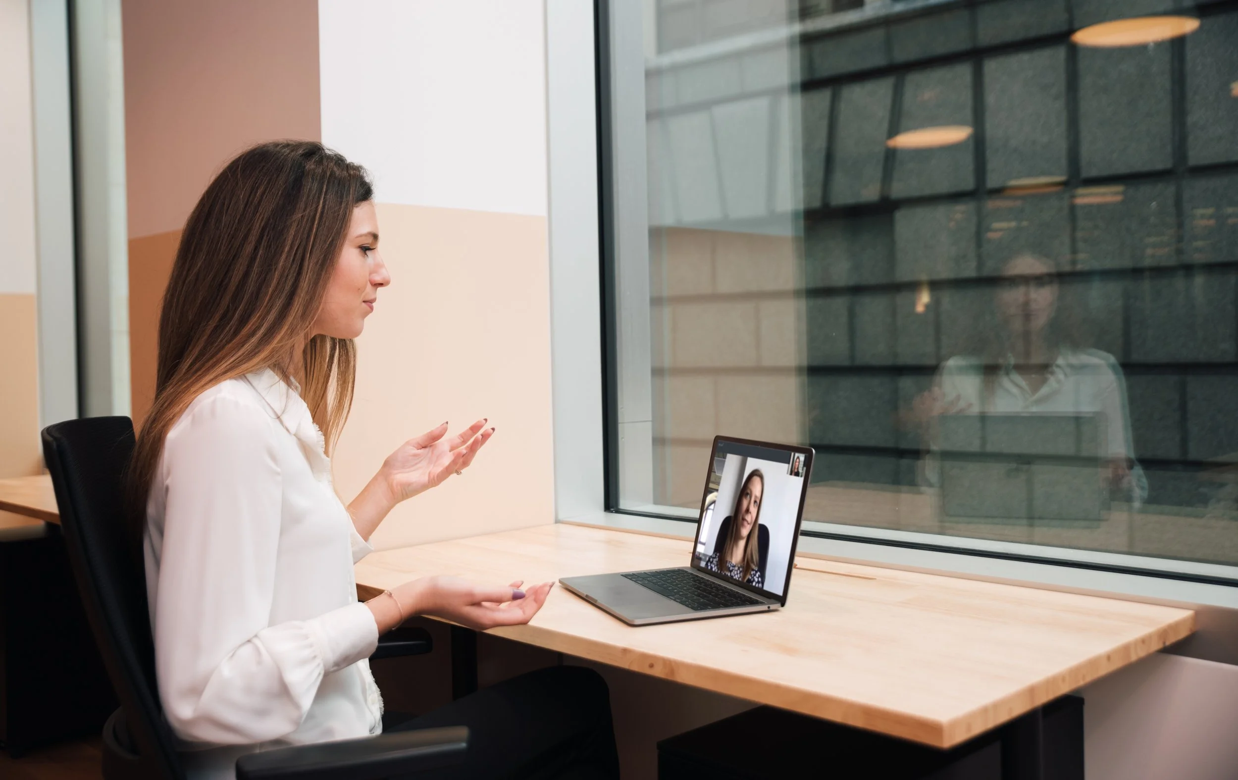 A woman participating in a video call with another woman, seen on her laptop screen, in an office setting by a window.