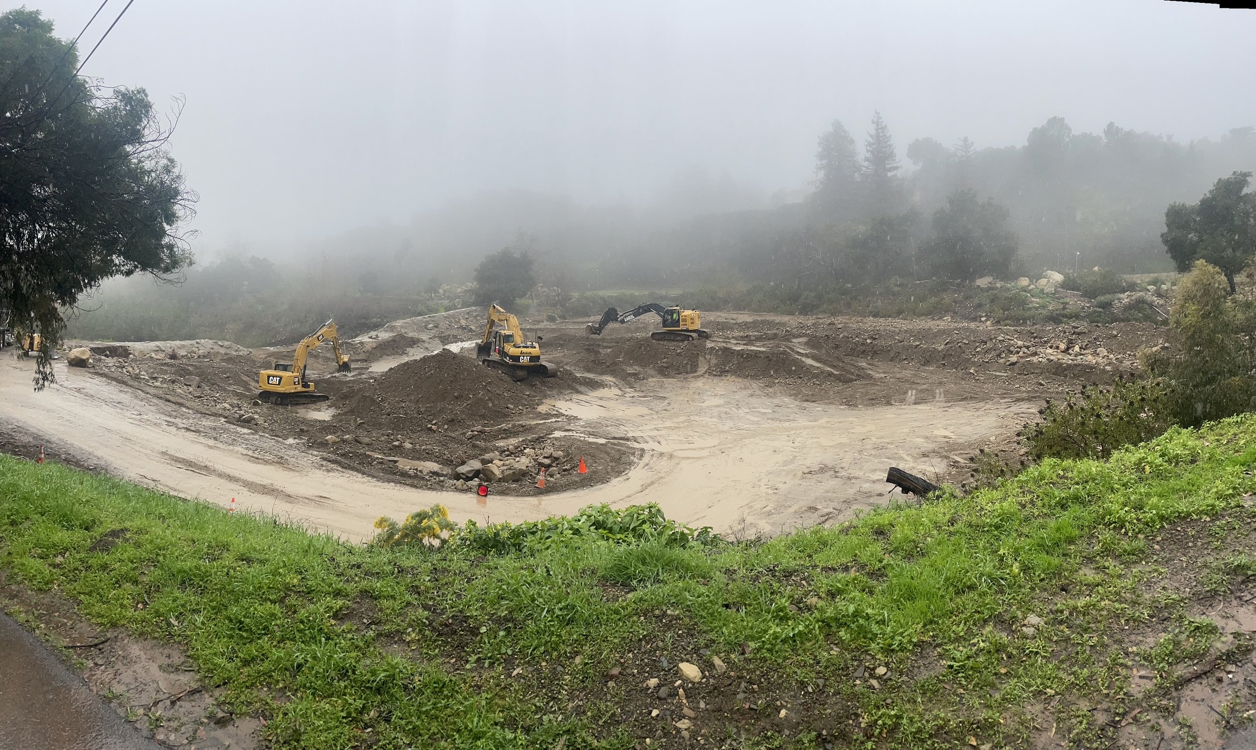 Construction site with multiple yellow excavators working on dirt excavation amidst foggy weather, surrounded by trees and greenery.