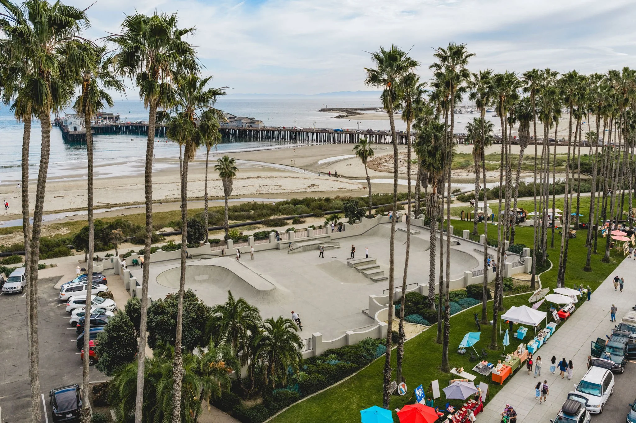 A view of a beachside park with palm trees, a skate park, and an outdoor market with tents, while the ocean and a pier extend into the water in the background.