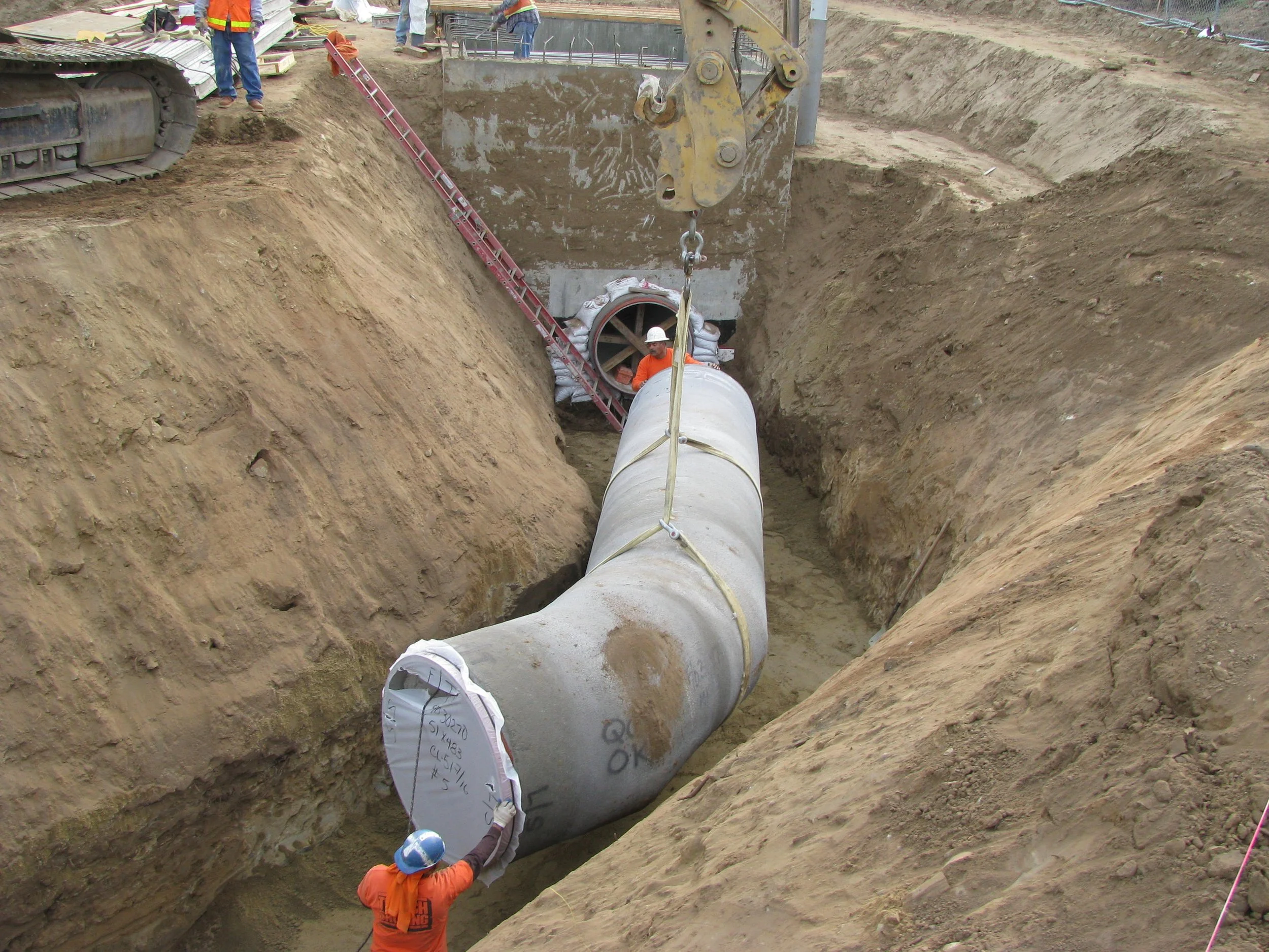 Construction workers are installing a large concrete pipe underground in a trench, with some workers visible on the sides and bottom of the trench, using equipment to position the pipe.
