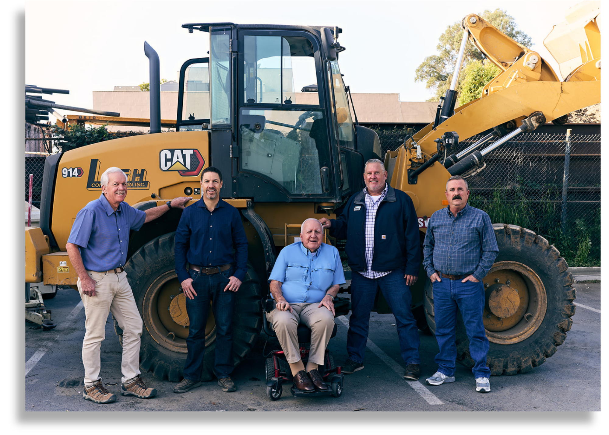 Group of six men posing in front of a large yellow construction vehicle at an outdoor lot, with trees and a building in the background.