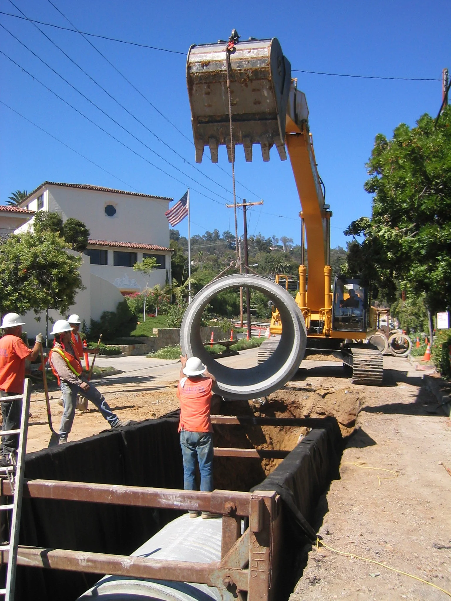 Construction workers are installing a large concrete pipe into a trench, with a yellow excavator lifting the pipe above the ground. The scene takes place outdoors on a clear, sunny day with a house, trees, and power lines in the background.