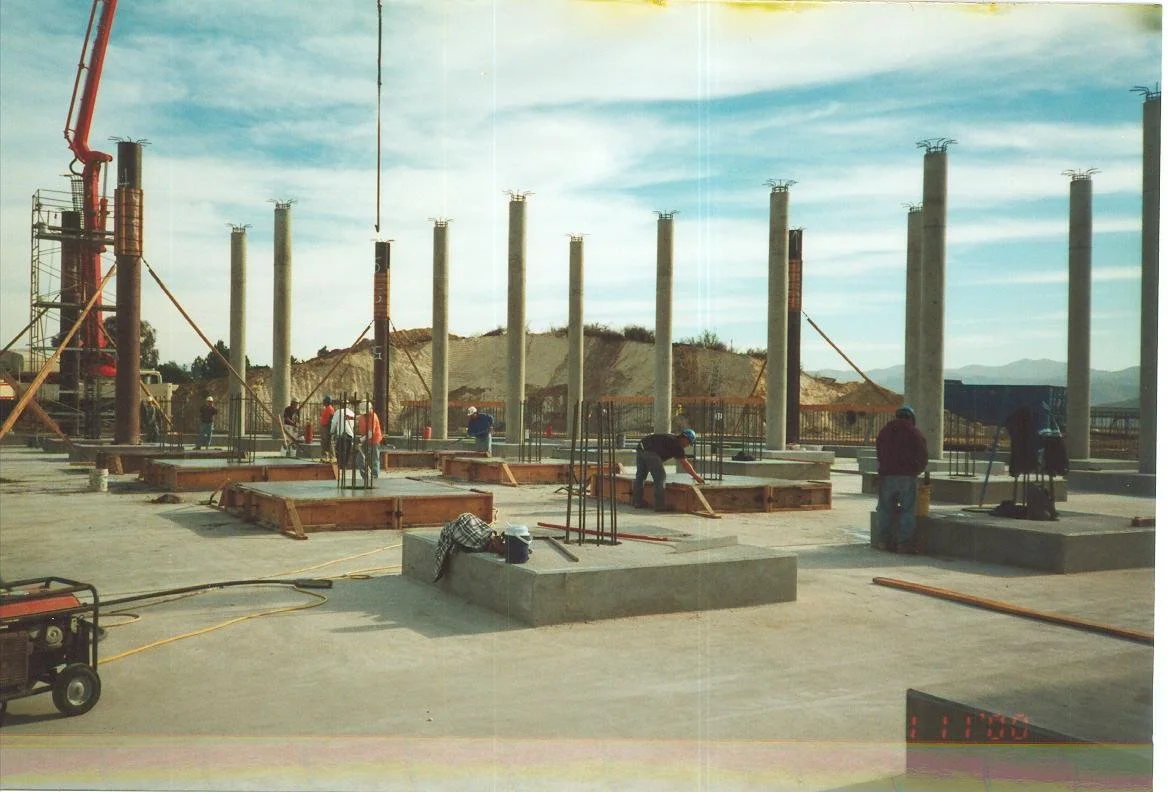 Construction site with workers building a structure with concrete and steel, supporting columns, and construction equipment under a partly cloudy sky.