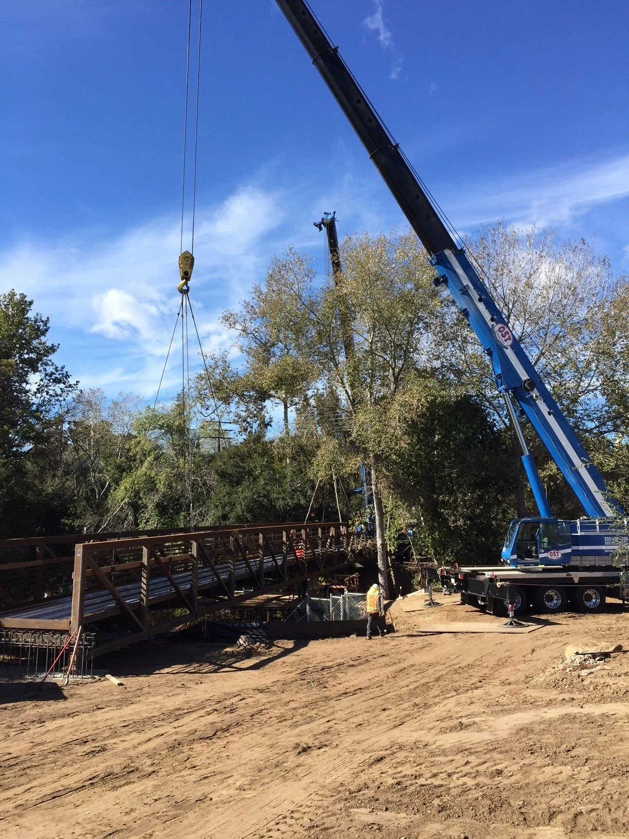 Construction workers using a large crane to build a bridge or similar structure outdoors, with trees and a blue sky in the background.